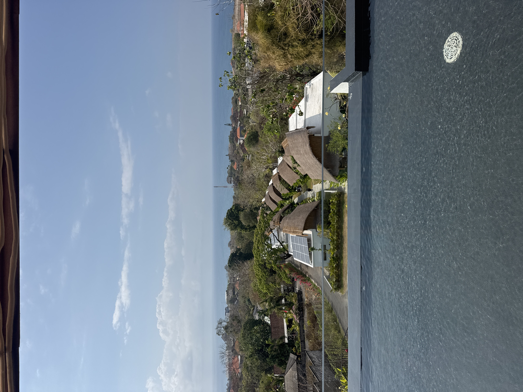 This is an eye-level shot taken from an infinity pool, looking out over a coastal landscape. The pool's dark, rippled water fills the foreground, with a decorative drain cover visible on the right. Beyond the pool's edge, a row of thatched-roof bungalows with white walls and solar panels sits nestled among lush greenery. A paved pathway runs alongside the bungalows, with a few pedestrians walking along it. In the middle ground, a mix of trees, houses with red tile roofs, and a telecommunications tower are visible. The background is dominated by the blue expanse of the ocean under a bright, partly cloudy sky. The scene suggests a resort or residential area in a tropical location. The geographical indicators suggest the location is Kuta Selatan, Indonesia.