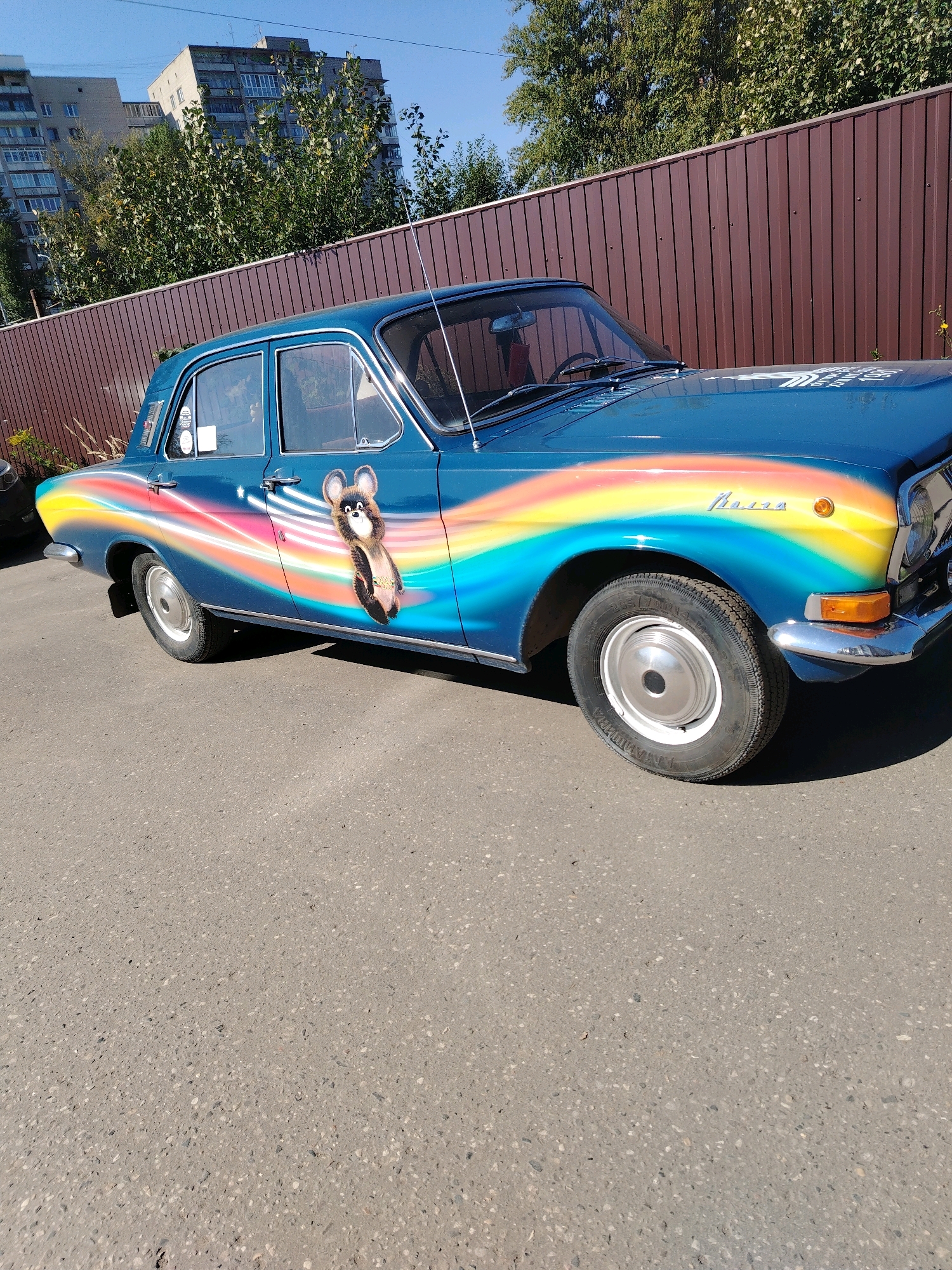 The image shows a blue vintage car with a rainbow decal and a cartoon character on its side, parked on an asphalt road in front of a brown corrugated metal fence. In the background, there are trees and apartment buildings. The car appears to be a GAZ-24 Volga. The specific location in Yaroslavl, Russia cannot be determined from the image alone.