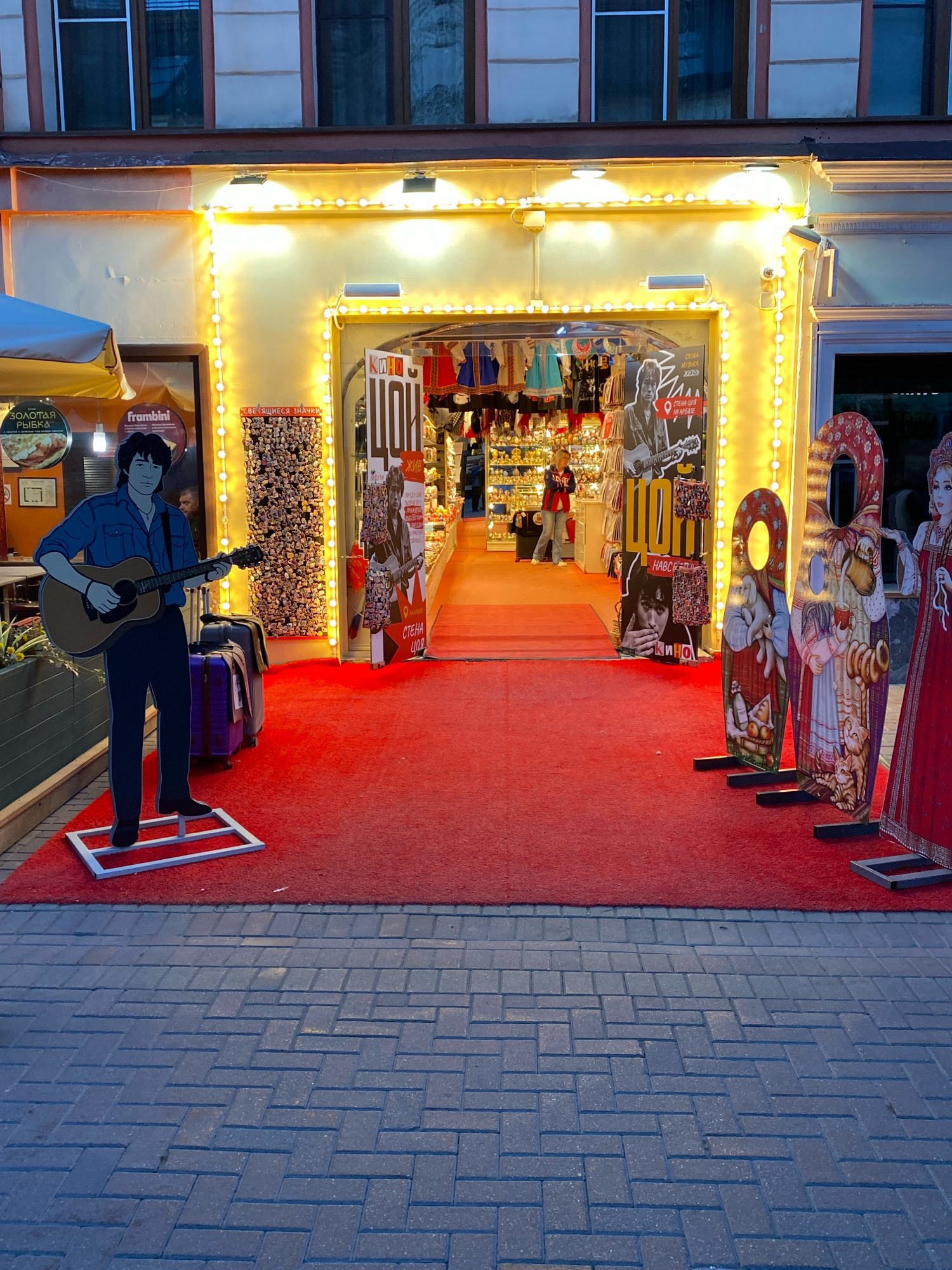 The image shows the entrance to a souvenir shop on a street in Moscow, Russia. The shop is decorated with bright yellow lights and a red carpet leads to the entrance. On the left side of the carpet, there is a life-sized cardboard cutout of a man playing a guitar, and two suitcases are placed behind him. Inside the shop, a woman can be seen browsing shelves filled with souvenirs. To the right of the entrance, there are several cardboard cutouts of women in traditional Russian dress, designed for photo opportunities. The street in front of the shop is paved with grey bricks. The overall scene suggests a lively and tourist-oriented area.