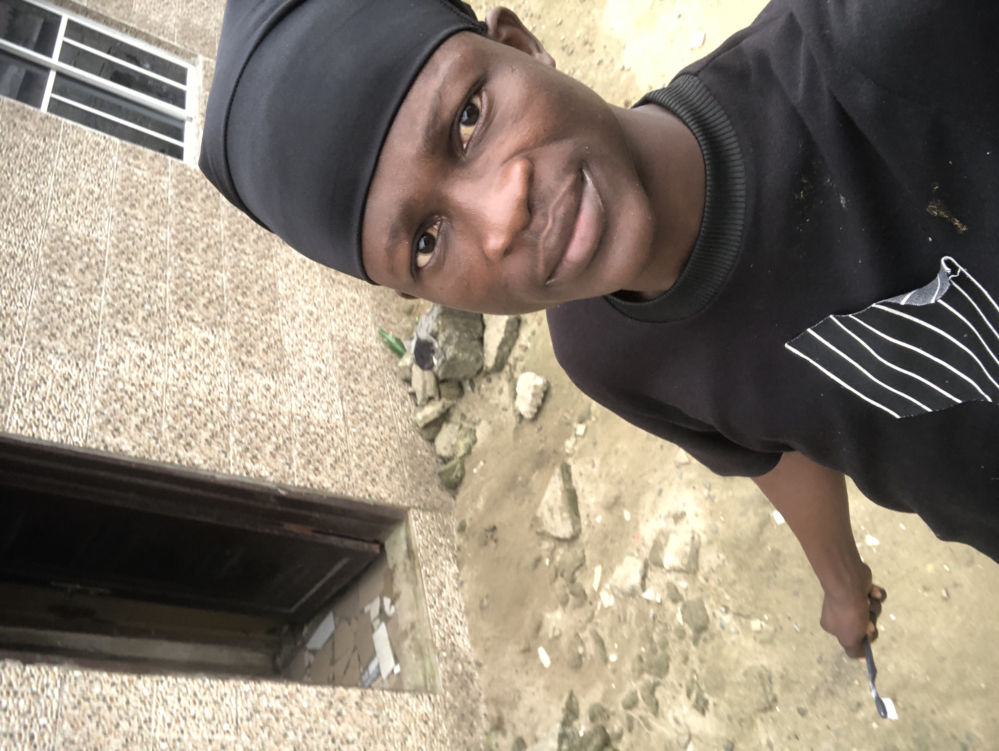 The image, taken in Ado, Nigeria, is a close-up, high-angle selfie of a young Black male. He is wearing a black beanie and a black long-sleeved shirt with white stripes on the left chest. He is holding a white object with a blue tip, possibly a toothbrush, in his right hand. The background consists of a beige, speckled building with a dark doorway and a barred window. The ground is composed of dirt, small stones, and debris, suggesting an outdoor, possibly unpaved area. The lighting is natural, casting soft shadows.