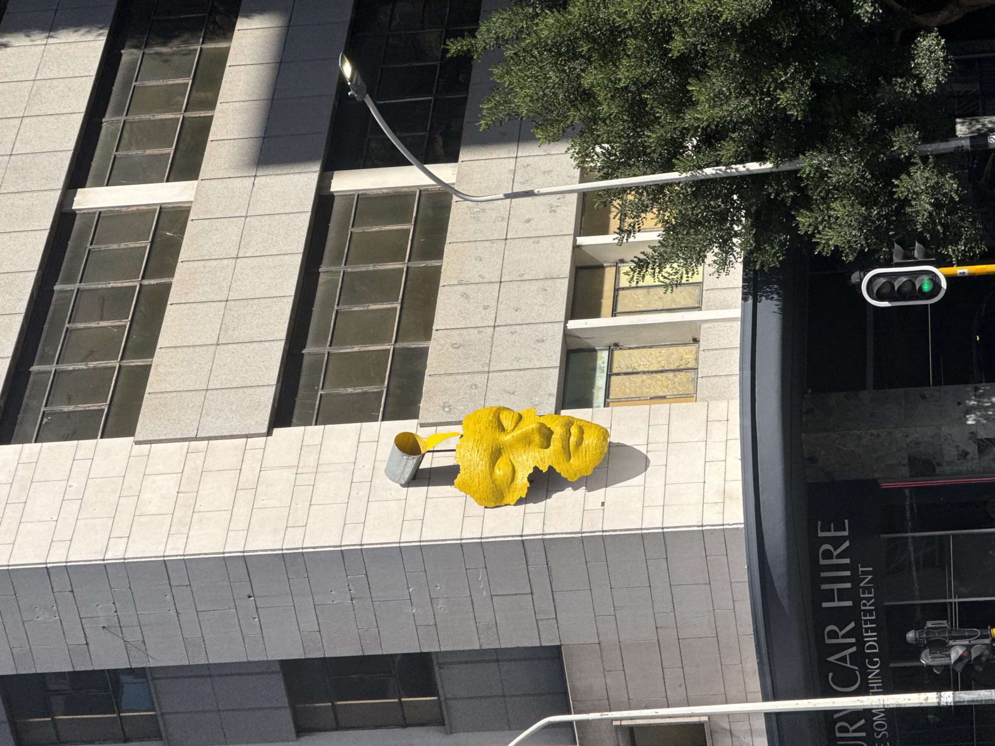 This image captures a multi-story building facade at 8 Riebeek Street, Cape Town, South Africa, under direct daylight. A prominent bright yellow, textured sculpture of a human head with closed eyes is affixed to the light-colored stone paneling. Above the sculpture, a grey metal bucket is mounted, angled to release a stream of yellow, viscous-appearing material onto the sculpture's head; this material matches the sculpture's color and texture. The building's facade is constructed from rectangular stone or concrete panels and features numerous dark-framed, multi-pane windows across its visible levels. A dark awning covers a lower commercial section, displaying partially visible white text on a black background that reads "URY CAR HIRE" and "E SOMETHING DIFFERENT." To the right, a white curved street light pole supports an LED luminaire, with its base partially obscured by dense green foliage. In the lower central portion, a traffic light on a yellow pole displays a green signal. Shadows from the sculpture are visible on the building's facade to its right, indicating sunlight from the left. No individuals are present in the scene.