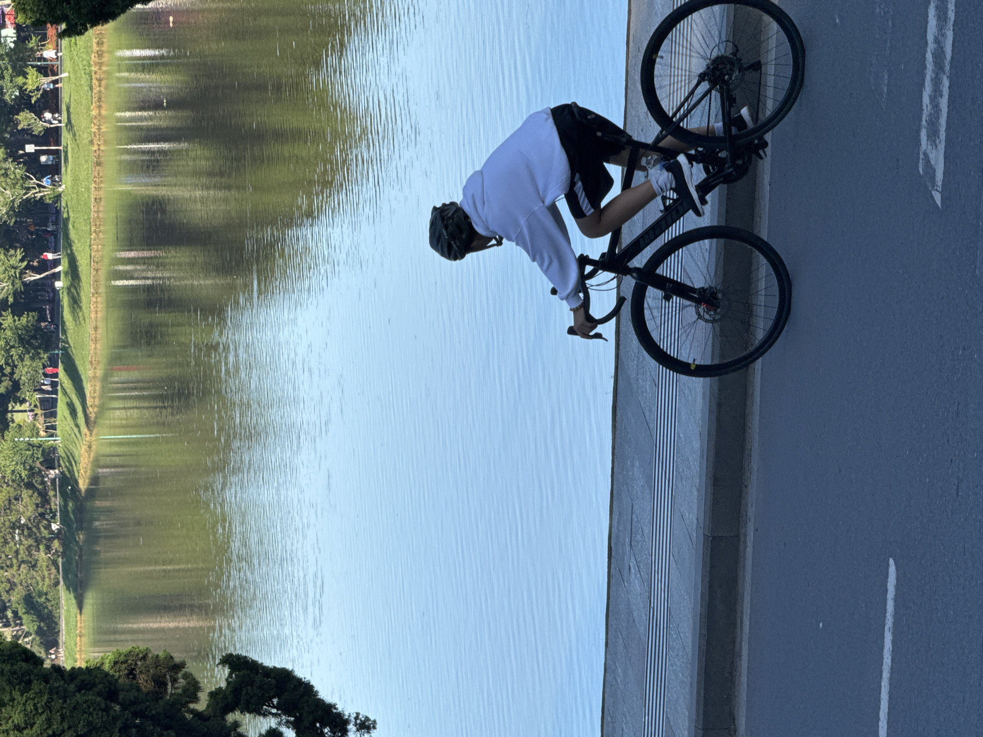 A person wearing a white hooded sweatshirt and black shorts rides a bicycle on a paved road next to a body of water. The person is also wearing a helmet. The water reflects trees and the sky. The road has a white line painted on it. The scene appears to be in Da Lat, Vietnam, as the style of the trees and the general atmosphere are consistent with the city's landscape. The overall impression is of a peaceful outdoor activity.