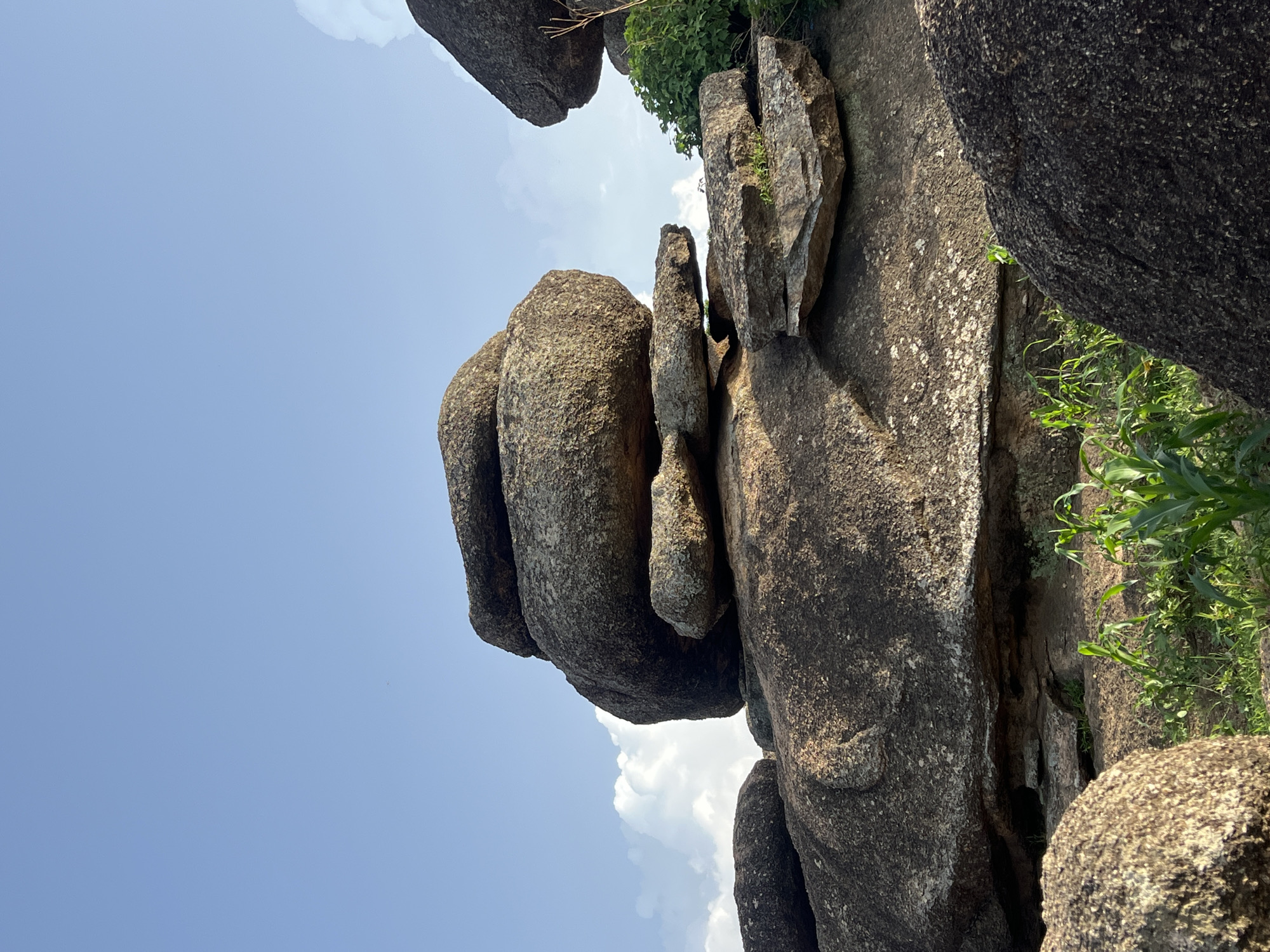 The media file captures a low-angle view of large rock formations against a bright blue sky with scattered white clouds. The rocks are weathered and appear to be part of a natural landscape, possibly a rocky outcrop or hillside. The foreground is dominated by the textured surfaces of these large stones, with some appearing to be stacked or precariously balanced. Green vegetation, including tall grasses or stalks, is visible in patches, particularly on the right side of the frame, suggesting an outdoor, natural environment. The sunlight indicates it is daytime, likely midday or afternoon, given the brightness of the sky. There is no discernible activity or event taking place, as the scene is static and focuses solely on the geological formations and surrounding nature. No people, text, or specific landmarks are visible to provide further contextual clues about the precise location within Jos, Nigeria, beyond the general impression of a rocky, vegetated outdoor area. The overall mood is one of natural grandeur and stillness.