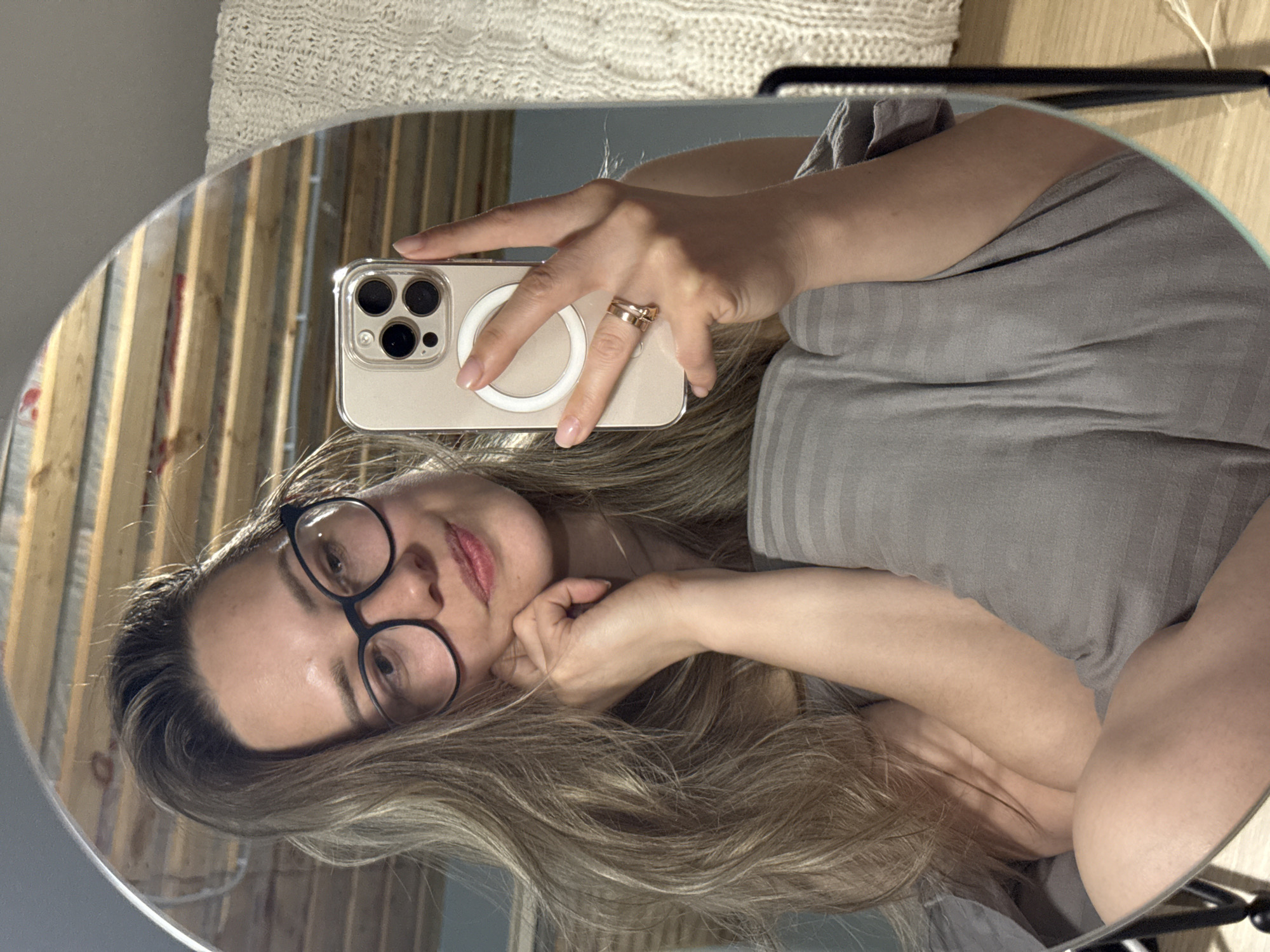 A person with long blonde hair and glasses is taking a selfie in a round mirror. The person is wearing a grey top and has a gold ring on their finger. They are holding a phone with a clear case and a white MagSafe charger ring on the back. The reflection shows a wall with wooden planks and a white knitted item in the background. The setting appears to be indoors, potentially a bedroom or living area. There is no information in the picture to indicate the location of Chaikovsky, Russia.