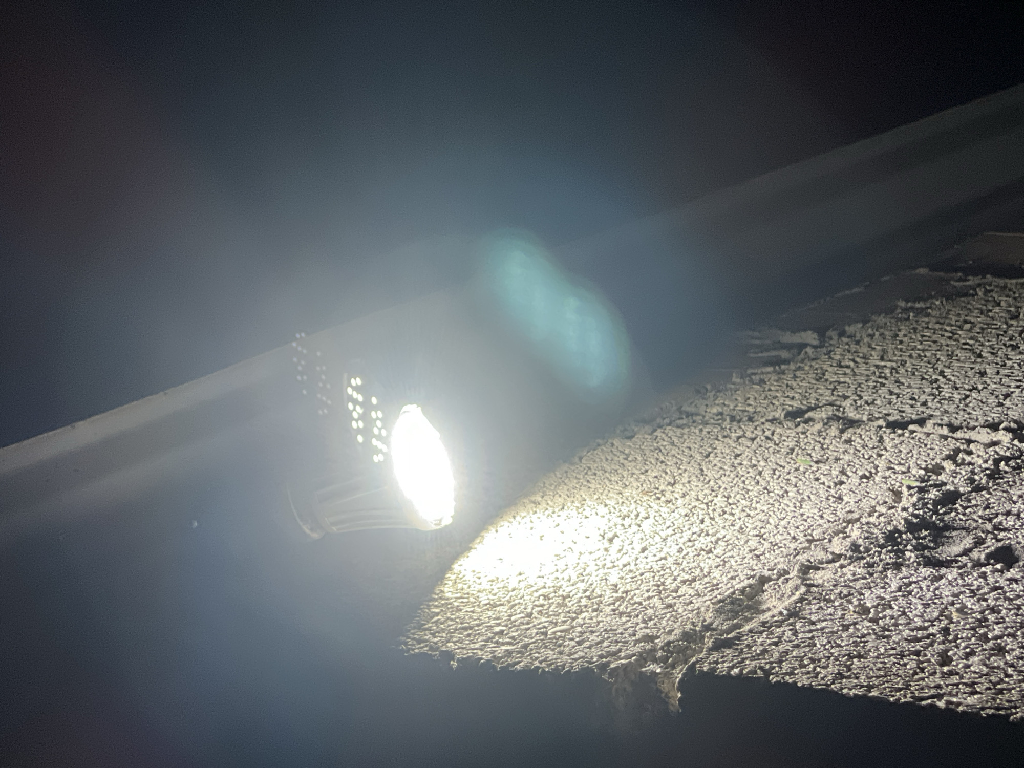 The image depicts an outdoor scene at night, illuminated by a bright overhead light source. The light, which appears to be an LED bulb, is positioned above and to the right of the frame. It casts a strong, white light onto a textured surface, likely a wall or structure, which occupies the lower left portion of the image. This textured surface is rough and uneven, with deep crevices and protrusions, creating a dramatic play of light and shadow.
To the right of the textured surface and slightly behind it, a smooth, angled object runs diagonally across the frame. This object is darker and appears to be part of a larger structure or architectural element. The background is dark and out of focus, suggesting an open, unlit area. A faint, greenish-blue lens flare is visible in the mid-right of the image, a common artifact of nighttime photography with bright light sources.
The image has a grainy quality, typical of low-light photography. There are no people or animals visible in the scene. The overall impression is one of an artificially lit, industrial or architectural space at night. Without further context, pinpointing the exact location in Komsomolskoye, Russia, is not possible from the visual information alone. However, the scene suggests an exterior wall of a building or outdoor structure.