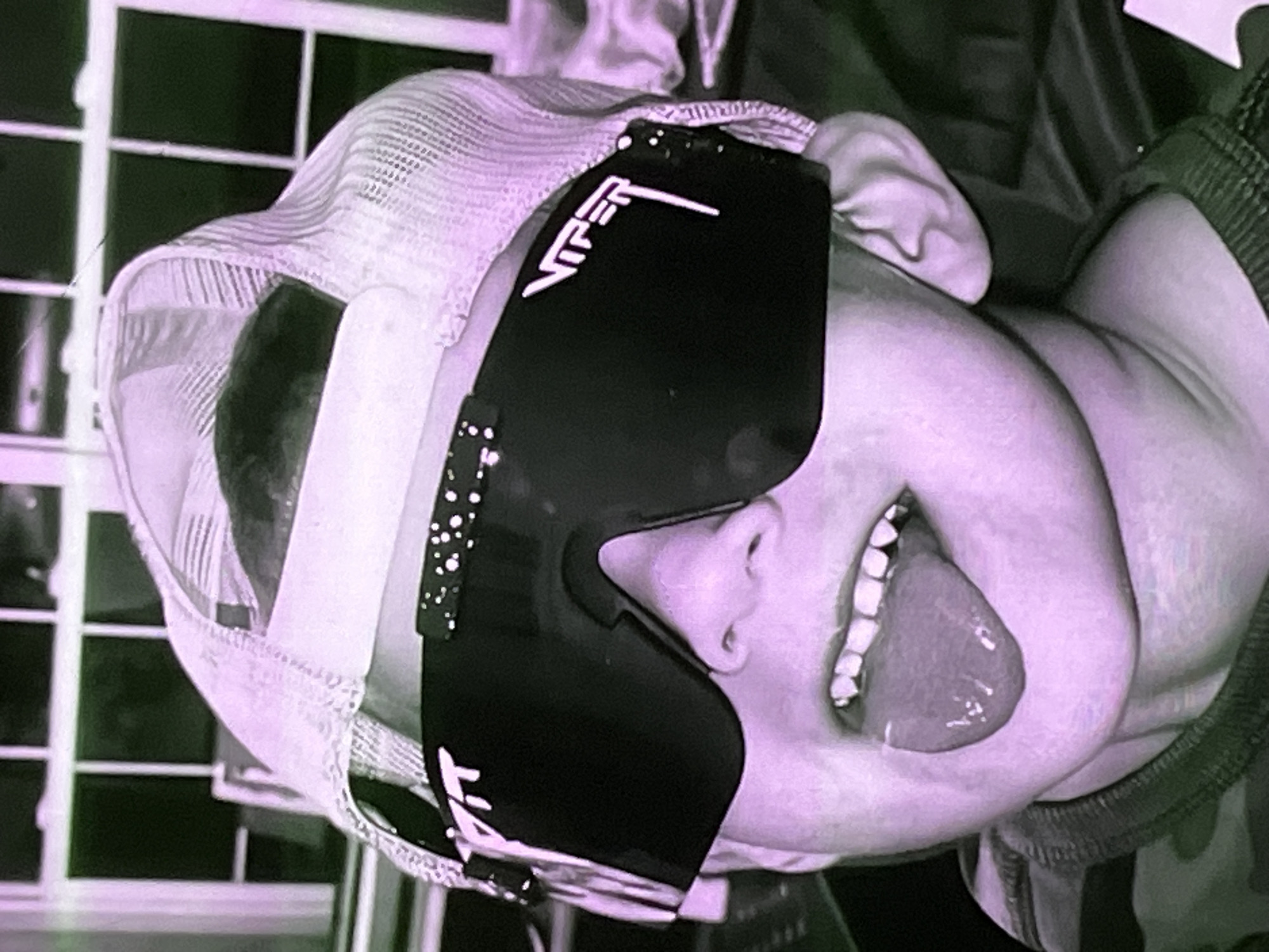 The image is a close-up, vertically oriented, low-resolution, digitally altered photograph of a young boy wearing sunglasses and a baseball cap. The boy is sticking his tongue out and his mouth is open, showing his teeth. The sunglasses are large and black with a white logo on the side. The baseball cap is light-colored and appears to be a trucker hat with a mesh back. The background is out of focus and appears to be a room with a window. The overall color tone of the image is purple and green, suggesting a filter or lighting effect. There is no information in the image to suggest the location is Cadek, Indonesia.