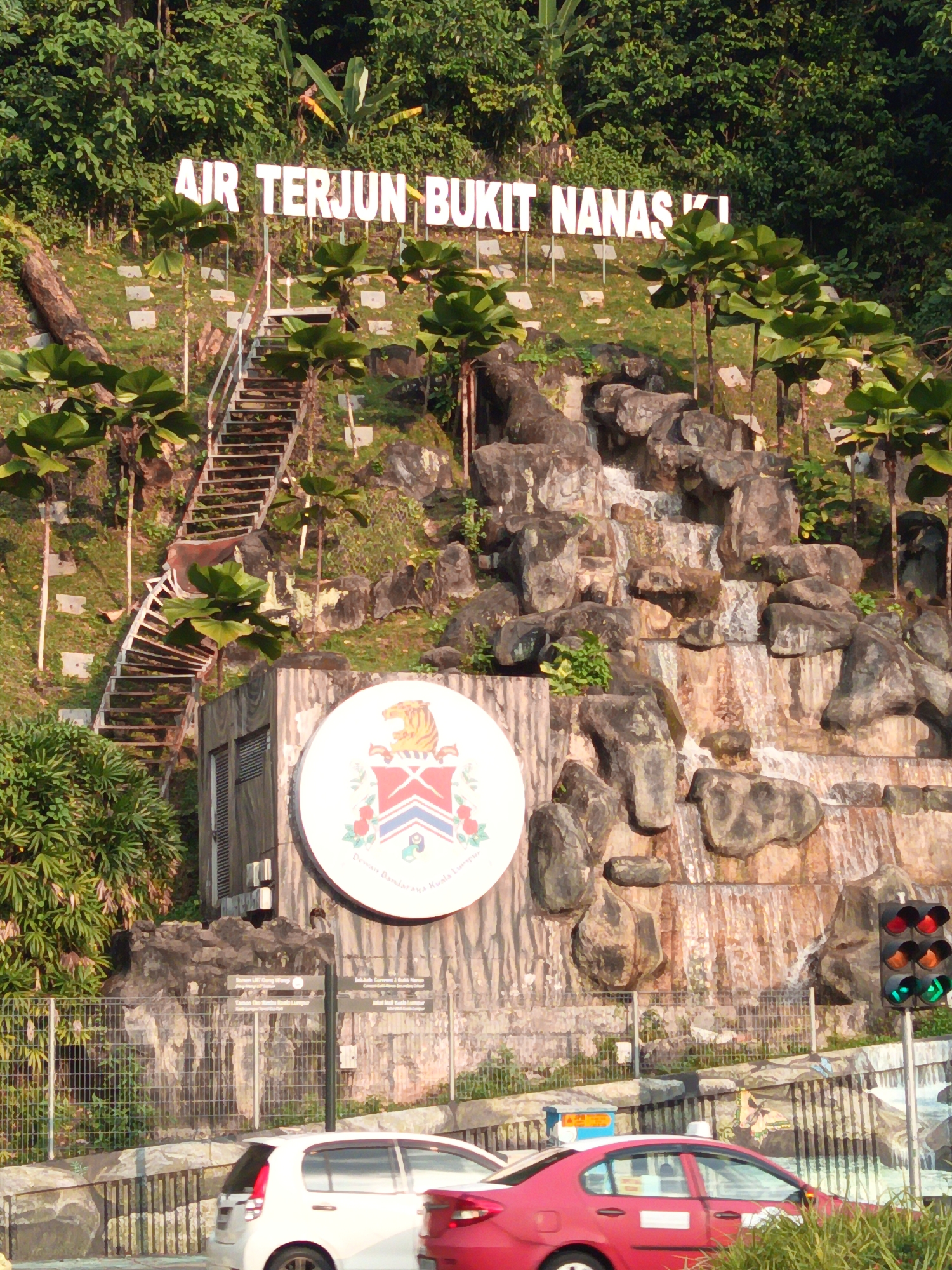 This image shows a tiered rock waterfall feature with a white sign in front of it reading "AIR TERJUN BUKIT NANAS KI". A staircase made of metal and wood ascends the left side of the waterfall. To the left of the waterfall is a grey concrete structure with a circular emblem on its front. The emblem is white with red and blue crests and an image of a tiger. The area is surrounded by lush green trees and vegetation. In the foreground, cars are visible on a road, and a traffic light is illuminated with green. This scene is located at the Bukit Nanas Waterfalls in Kuala Lumpur, Malaysia, a historical forest reserve in the city center.