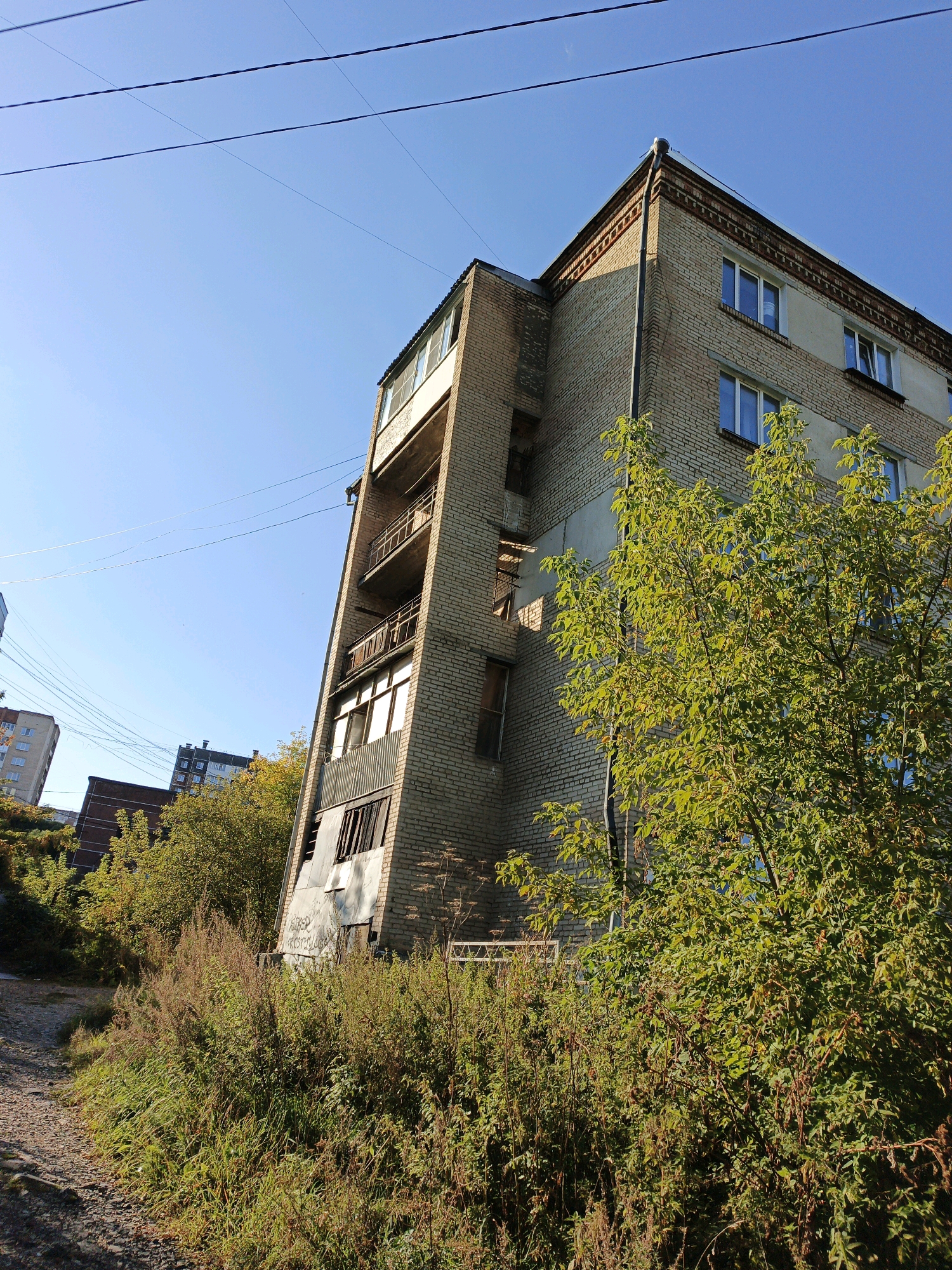 This is a low-angle, full shot, outdoor photograph taken in Zlatoust, Russia, on a clear, sunny day. The image features a multi-story brick building with a unique architectural design. The building is constructed with light-colored brick and has several windows and balconies. The building's facade slopes outwards from top to bottom on the right side of the image. In the foreground, lush green and brown vegetation, including tall grasses and trees, partially obscures the building. A gravel path is visible on the left side of the image, leading away from the viewer. Overhead, multiple power lines crisscross the bright blue sky. In the background, other buildings and trees are visible. The scene appears to be an urban residential area. There are no people present in the image.
