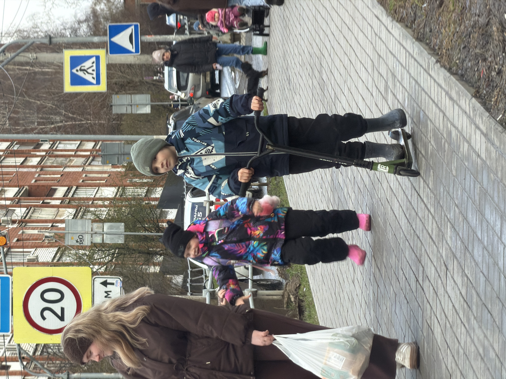 The image depicts a daytime scene on an urban sidewalk in Izhevsk, Russia, under overcast skies. The ground is paved with light grey interlocking bricks, and leafless trees in the background suggest late autumn, winter, or early spring.
In the foreground, a young boy, dressed in a blue and white patterned winter jacket with "FUNJIANG" visible on the sleeve, dark pants, and a grey knit hat, rides a green and black "MICRO" brand scooter. He wears dark grey boots. Standing near him is a younger child, likely a girl, wearing a vibrant multi-colored winter jacket, dark pants, and a black knit hat, with visible pink socks or footwear.
Behind them, a woman with long blonde hair, donning a brown jacket and a dark brown knit headband, walks while carrying a white plastic shopping bag. Further back, another adult in a dark jacket and blue jeans, wearing bright green boots, is visible, possibly with a stroller and child. Other pedestrians and vehicles are seen in the distance.
Several road signs are prominent: a yellow square sign indicating a pedestrian crossing, a blue square sign with an inverted white triangle (possibly for a hump or dip), a yellow circular speed limit sign showing "20", and a white rectangular sign with an upward arrow and "M↑" (indicating meters). Brick apartment buildings line the street, and an amber traffic light is also visible. A vehicle in the background has "РАНЫ 55" partially visible on its side. The scene portrays a casual outing with children at play on a sidewalk.