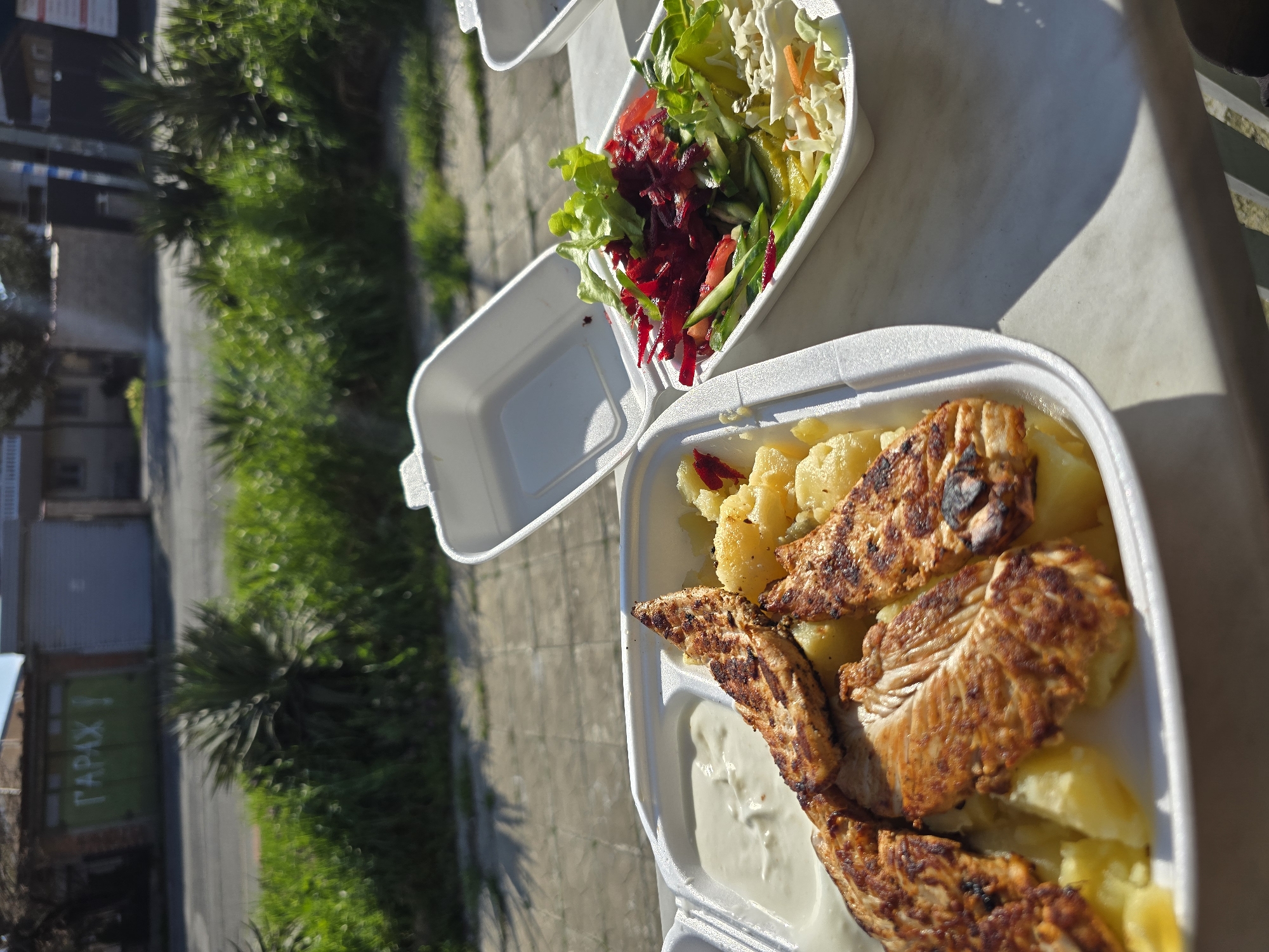 The image shows a takeout meal consisting of grilled chicken, boiled potatoes, tzatziki sauce, and a salad in white styrofoam containers. The meal is placed on a white surface, likely a table. In the background, blurred, is an urban scene. The scene includes a green-painted garage, vegetation, a street, and buildings. The scene is most likely in Burgas, Bulgaria.