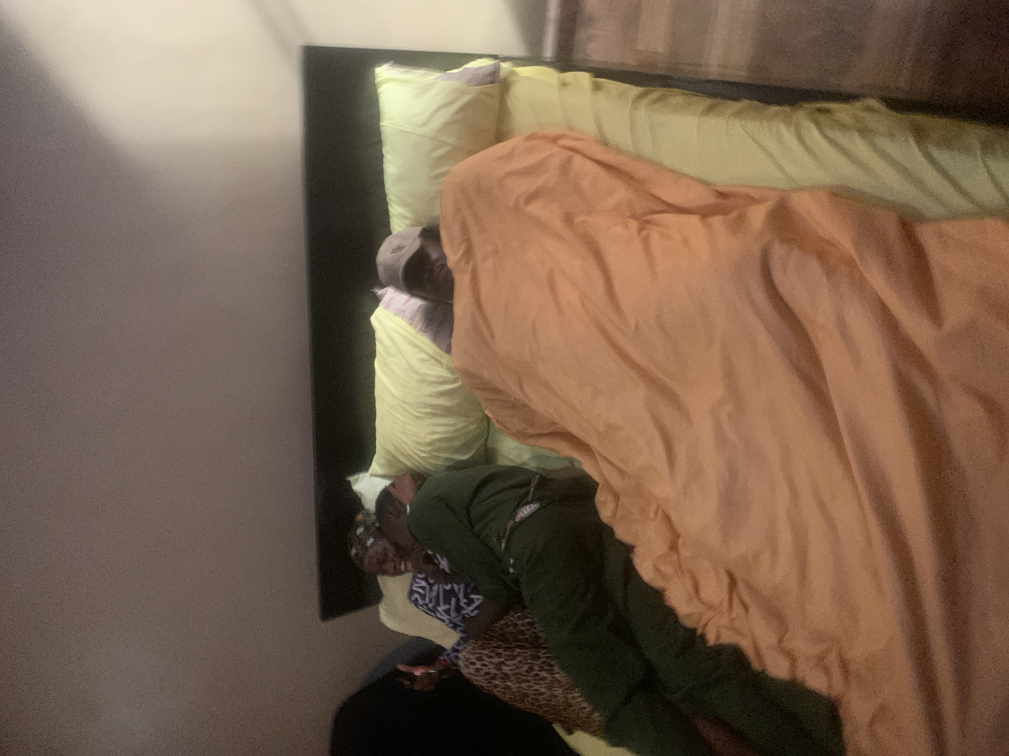 Two individuals are lying in a bed. The person on the left, wearing a headscarf and a patterned top, is smiling and embracing the person next to them. This person is wearing a dark green tracksuit. The second individual, on the right, is wearing a baseball cap and is mostly covered by an orange blanket. The bed has yellow sheets and pillows. The scene appears to be indoors, likely a bedroom, given the presence of a bed and a bedside table to the right of the image. The lighting is dim, suggesting it may be evening or night. This scene is located in Nkpor, Nigeria.
