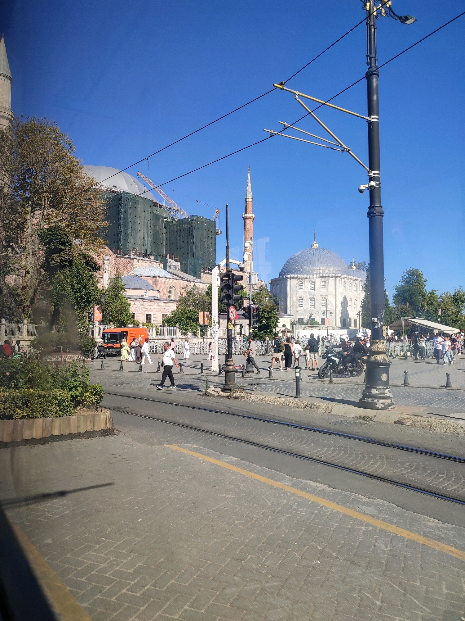 The image shows a street scene in Sultanahmet Square, Istanbul, Turkey, with the Hagia Sophia complex visible in the background. The complex's main dome is partially covered by green scaffolding, and a crane is present. To the right of the Hagia Sophia, another large building with a dome, possibly the Blue Mosque, is visible, also appearing to be undergoing renovation with scaffolding. The square is paved with cobblestones, and tram tracks run through the foreground. Several people are walking and crossing the street, including a man in a white shirt and dark pants walking across the tram tracks. An orange utility vehicle is parked on the side of the street. A tall lamppost with security cameras and overhead tram wires is prominently featured on the right side of the image. The sky is clear and blue.