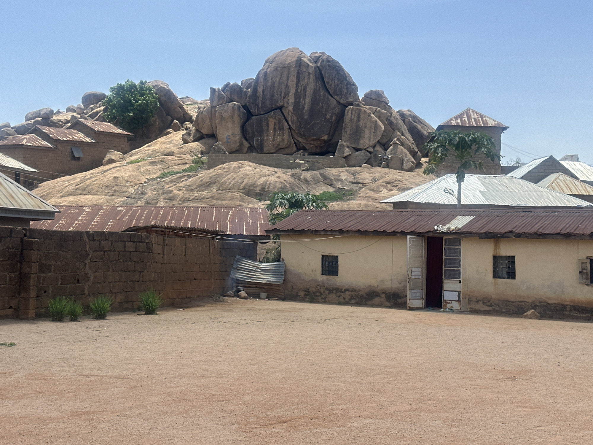 The scene depicts a rural area, likely in Jos, Nigeria, based on architectural style and terrain. The foreground is a dirt courtyard. The midground features a wall made of brown bricks, and a building with a corrugated metal roof and off-white walls. An open doorway reveals a dark red interior. Several windows with bars are visible on the building. A makeshift shelter constructed from corrugated metal is present between the wall and the building. In the background, a large rocky outcrop with boulders is visible. There are structures built into the rocks, some with corrugated metal roofs. The sky is a clear, light blue.