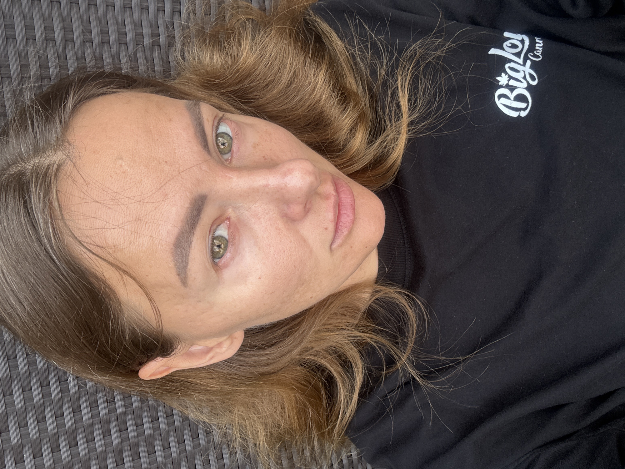 A woman with light skin and light brown hair is lying on a textured grey surface. She is wearing a black long-sleeved shirt with white lettering that includes the word "Biglo". Her eyes are green. The location is unidentifiable; there is no evidence in the image to indicate it is North Kuta, Indonesia.
