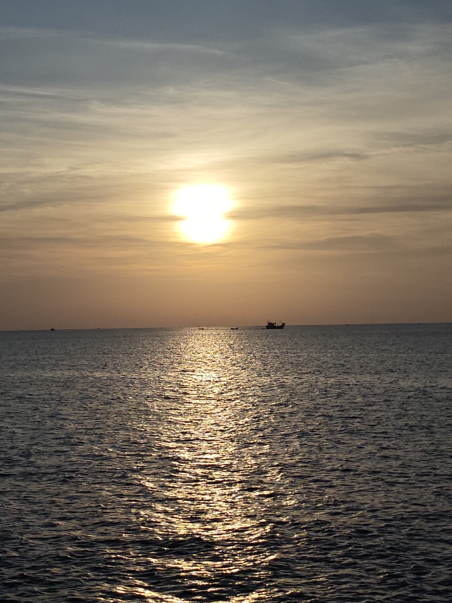 The image depicts a sunset scene in Lhokseumawe, Indonesia. The sun is positioned near the horizon, casting a bright glow on the water surface. The water is dark and choppy, with sunlight reflecting off the waves. A small boat can be seen in the distance, silhouetted against the bright sky. The sky is light in color, with some cloud cover near the top of the image. There are no visible people or other objects in the scene.