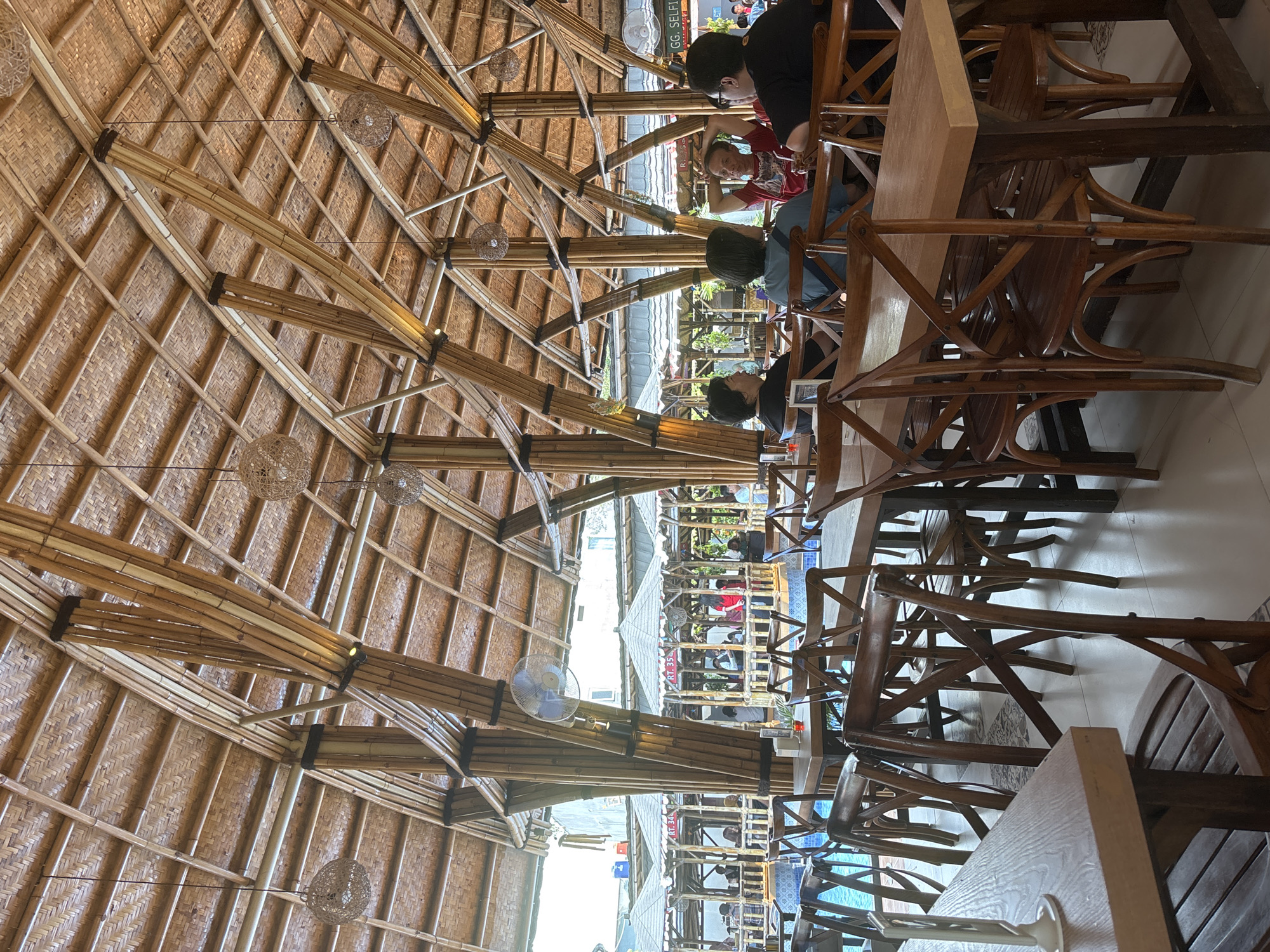 This image depicts the interior of a restaurant in West Jakarta, Indonesia, featuring a distinctive bamboo architectural design. The ceiling is a complex lattice of bamboo poles, creating a natural and rustic aesthetic, with woven bamboo panels forming the roof surface. Several spherical pendant lights hang from the ceiling. The dining area is furnished with wooden tables and chairs, many of which have an X-shaped backrest.
In the background, there are separate gazebo-like dining areas, also constructed with bamboo, situated around a swimming pool. People are visible seated in these gazebos, engaged in dining and conversation. The overall atmosphere suggests a casual and open-air dining experience. A ceiling fan is mounted on one of the large bamboo support pillars. The flooring appears to be light-colored tiles. The restaurant likely offers a view of the pool, adding to its ambiance.