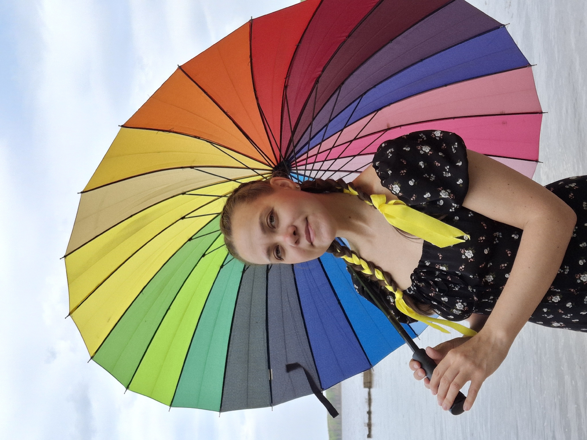 A medium shot depicts a young woman holding a large, multicolored umbrella. The umbrella features sections of red, orange, yellow, green, blue, purple, and pink. The woman has light skin, brown hair styled in braids with yellow ribbons, and is wearing a black dress with a floral pattern. She holds the black handle of the umbrella with her right hand. The background consists of a cloudy sky and a body of water. A concrete structure is visible on the horizon near the waterline. The location is Unknown, Unknown.