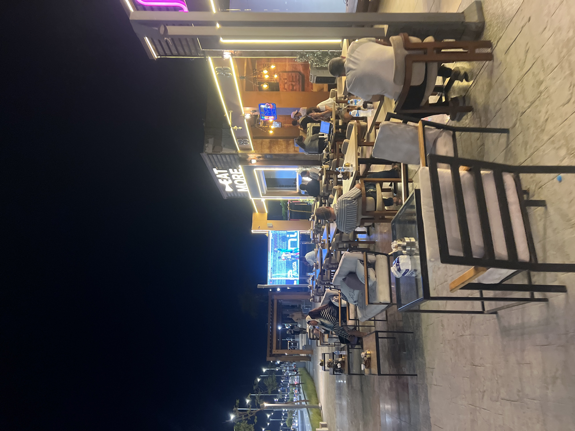 This image depicts an outdoor seating area of a cafe or restaurant at night in Sharm El Sheikh, Egypt. The scene is illuminated by artificial lights, including string lights, spotlights, and a large digital screen displaying a sports event. In the foreground, several tables and chairs are arranged on a paved patio.
Several patrons are seated at the tables, engaged in conversation or observing the screen. Some are eating or drinking. The cafe itself has a modern facade with signage that reads "EAT MORE." The background shows a street with lampposts and trees, indicating a well-developed urban area. The overall atmosphere appears relaxed and social, typical of a dining establishment in the evening.