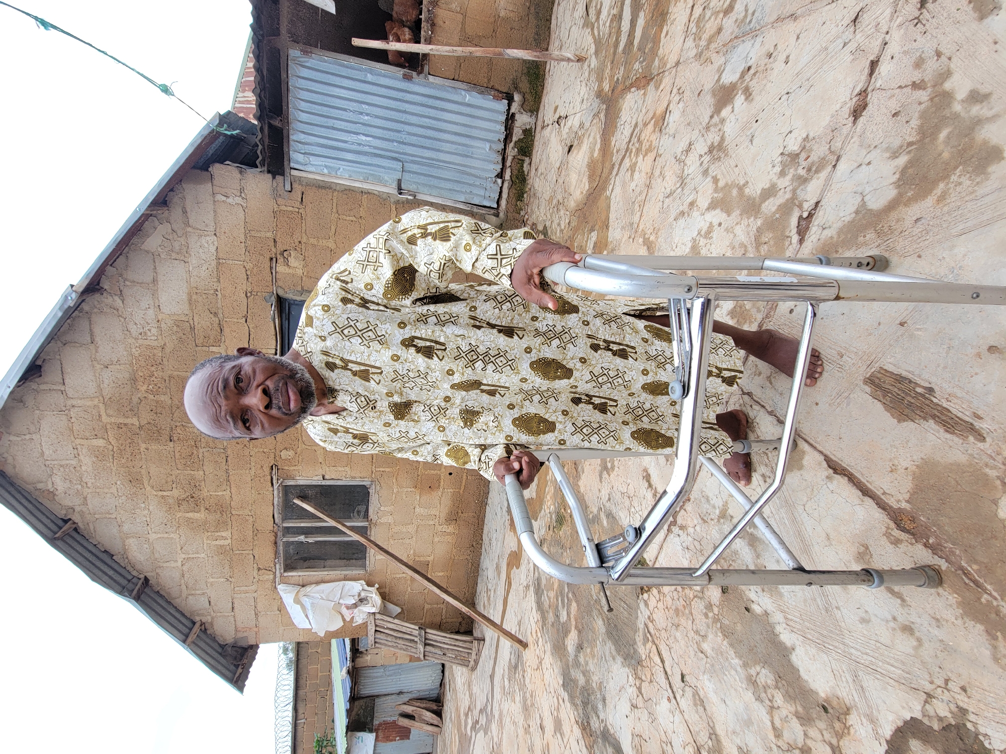 An elderly Black man stands outdoors, supported by a walker. He is wearing a light-colored, long-sleeved shirt with a repeating pattern of figures and geometric shapes. His bare feet are visible, and he has a beard and bald head. The man is positioned in front of a building constructed of light brown bricks with a corrugated metal roof. To his left, there is a window covered with a piece of white fabric and a wooden plank leaning against the building. To his right, a metal door is visible. The ground is a concrete surface with water stains and cracks. The overall environment appears to be rural or a less developed urban area, given the rustic nature of the building and the ground.