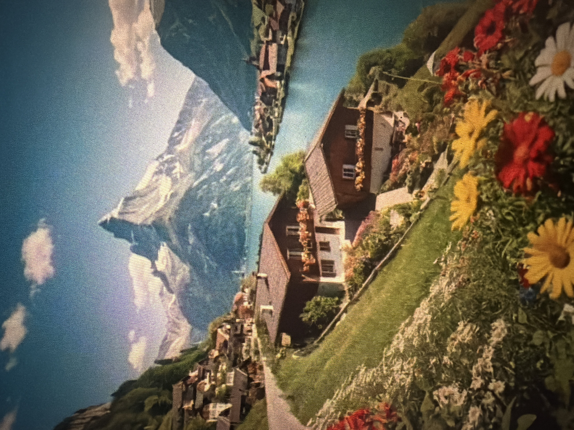 The image depicts a picturesque mountain village scene with a prominent snow-capped peak in the background. In the foreground, a hillside is adorned with vibrant flowers and lush green grass, leading to traditional wooden houses with red roofs. A winding road snakes up the hill, passing by these houses. Further in the distance, a serene blue lake is visible, with a town or village nestled along its shores. The sky is a clear blue with a few wispy clouds. The scene evokes a sense of tranquility and natural beauty, characteristic of a European alpine setting. The image is a photograph, possibly a painting or digitally enhanced image due to its stylized appearance and vibrant colors. There are no people visible in the image.