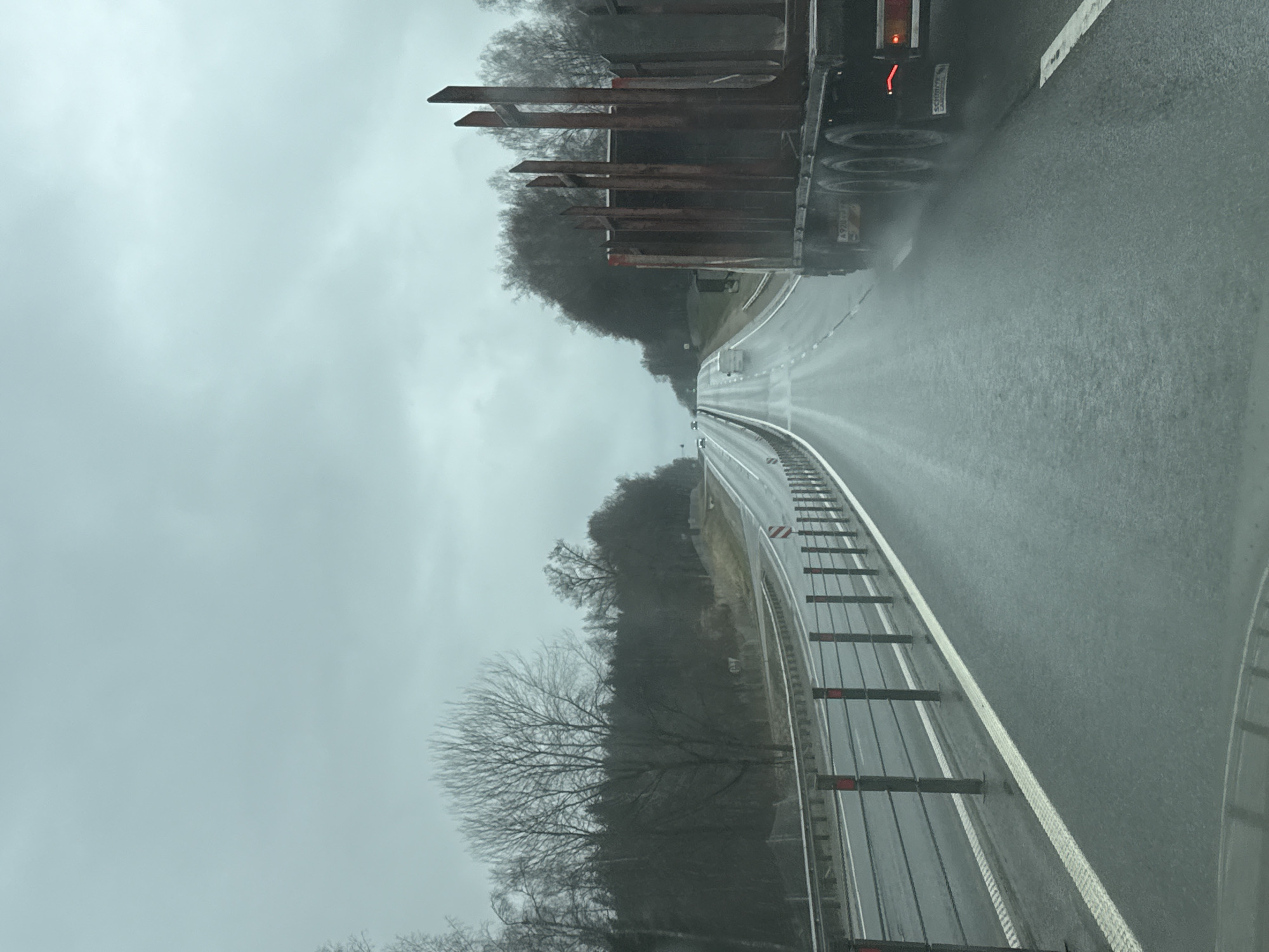 A view from a vehicle traveling on a highway under rainy conditions. To the right, a guardrail with white and red reflectors runs along the edge of the road. On the left side of the road, a vehicle, possibly a truck, is moving in the same direction. The sky is overcast and gray. Trees line both sides of the highway. There is no indication in the image that this is in Korobovo, Russia.