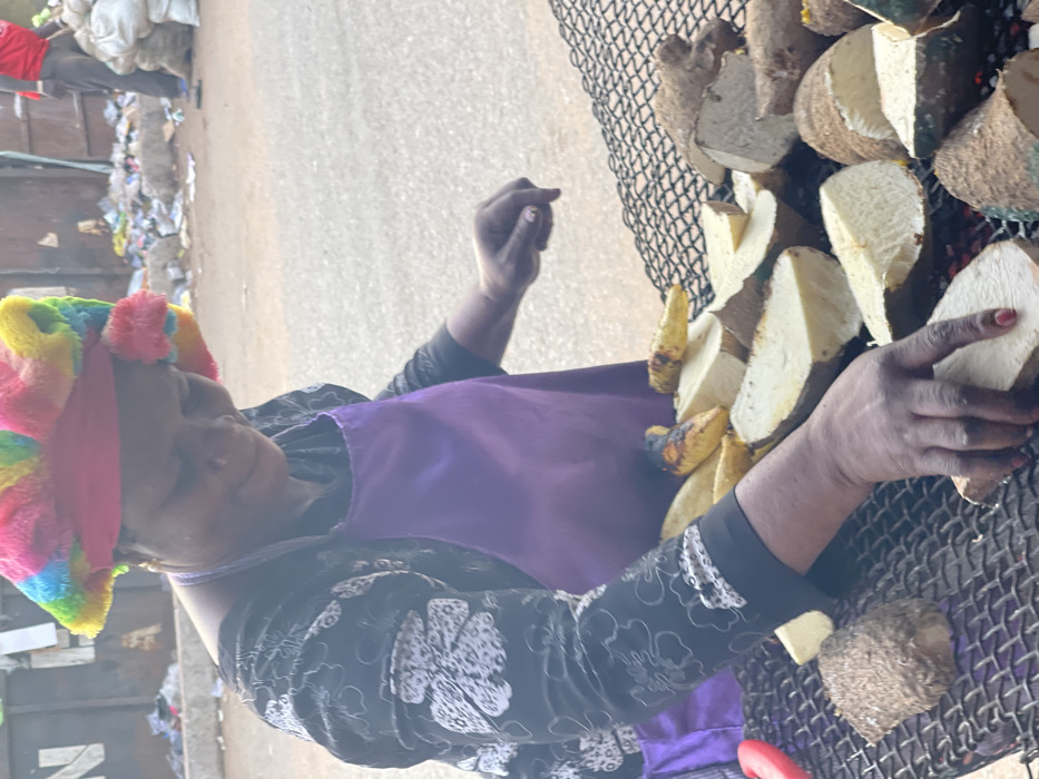 A person with a colorful headscarf and a purple apron is tending to a grill. The grill is covered with halved and quartered pieces of yams and plantains, which appear to be roasting or grilling. The person’s hands are visible as they handle the food on the grill. The setting appears to be outdoors, possibly a street scene, with a blurry background of buildings and debris.
