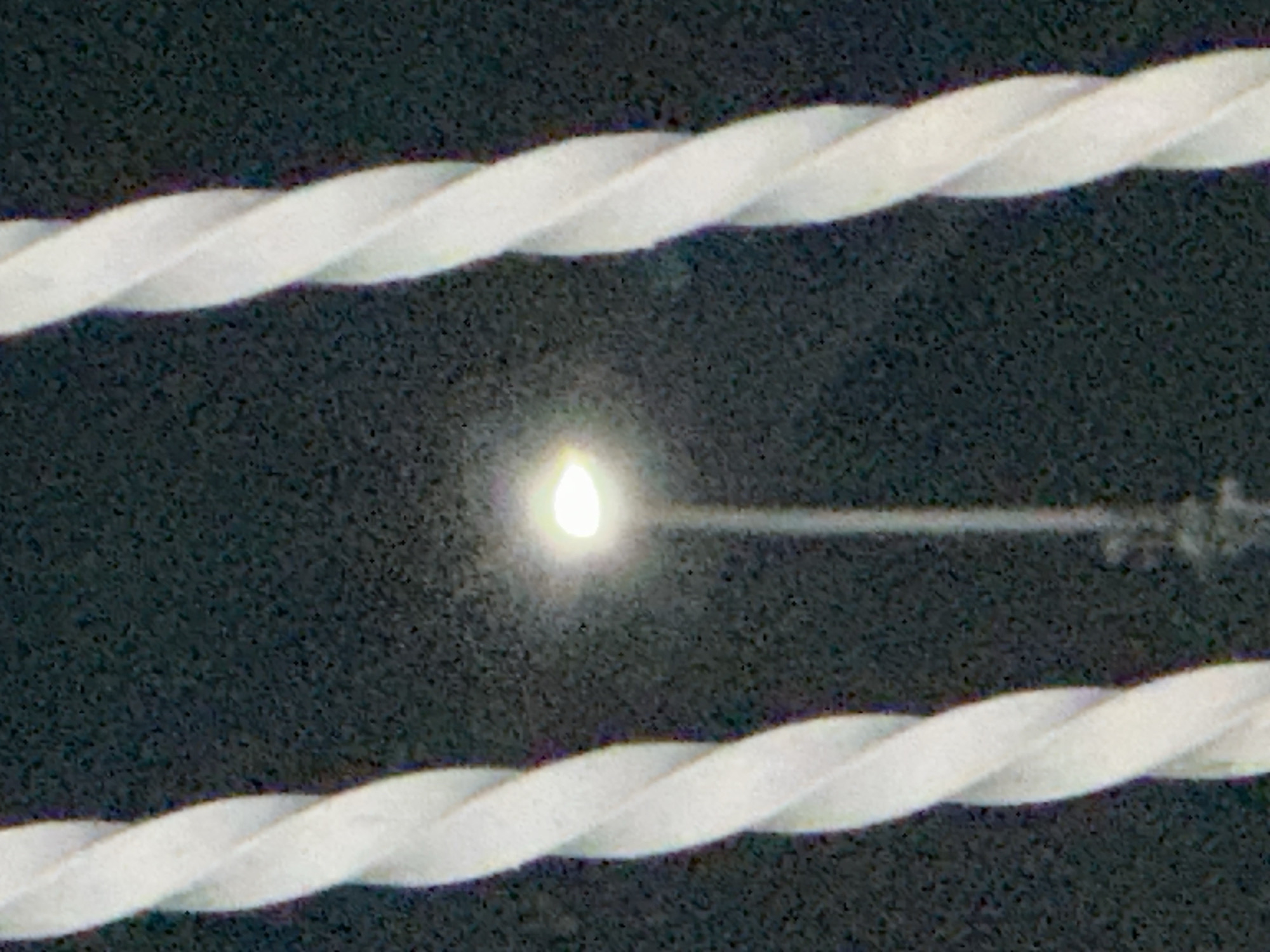 In El Maco, Venezuela, a streetlamp with a bright light is visible behind two twisted white poles. The background is dark and appears to be nighttime. The image is grainy, suggesting it was taken with low light or a low-quality camera.