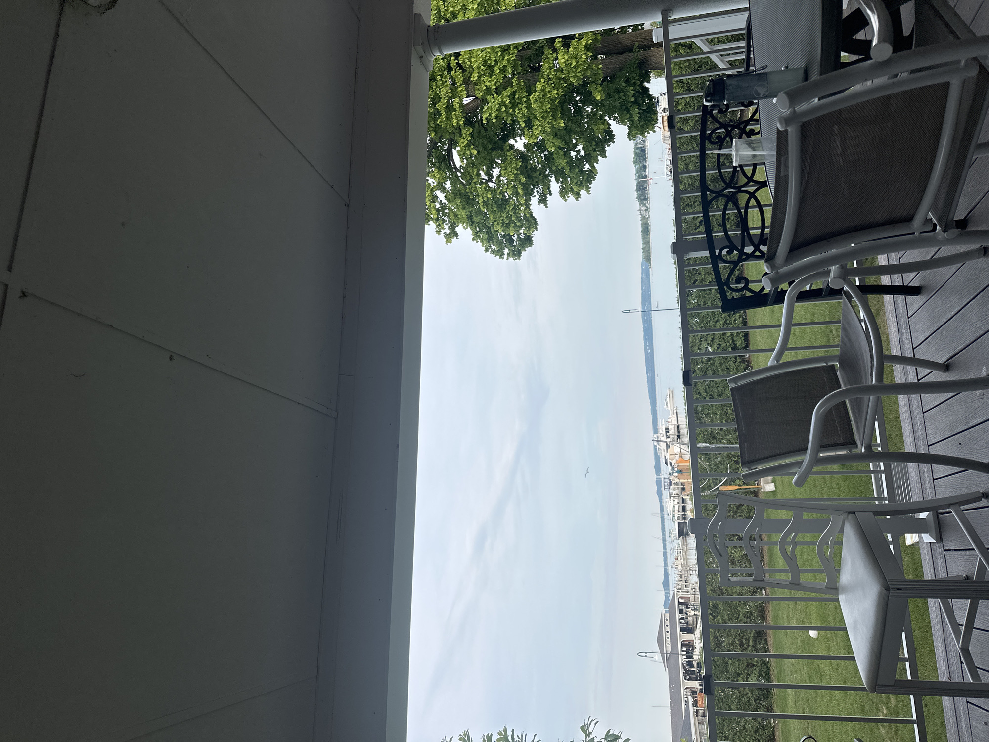 The image captures a view from a covered porch or deck overlooking a body of water, likely a harbor, in Harbor Springs, Michigan, United States of America. In the foreground, several outdoor chairs and a table are visible, suggesting a seating area. The chairs vary in design, with one white, ornate-backed chair and other more contemporary, lighter-colored chairs. A dark, patterned chair is also present. On the table, a clear plastic cup and a water bottle are placed.
Beyond the seating area, a metal railing separates the deck from a green lawn. In the middle ground, a line of docked boats, predominantly white, are visible along a waterfront with buildings in the background. The water extends to the horizon, where it meets a hazy sky with scattered clouds. To the right, a large, leafy green tree with a thick trunk is prominent, with a white pillar of the porch visible beside it. The overall scene depicts a relaxed outdoor setting with a view of a marina and a calm body of water.