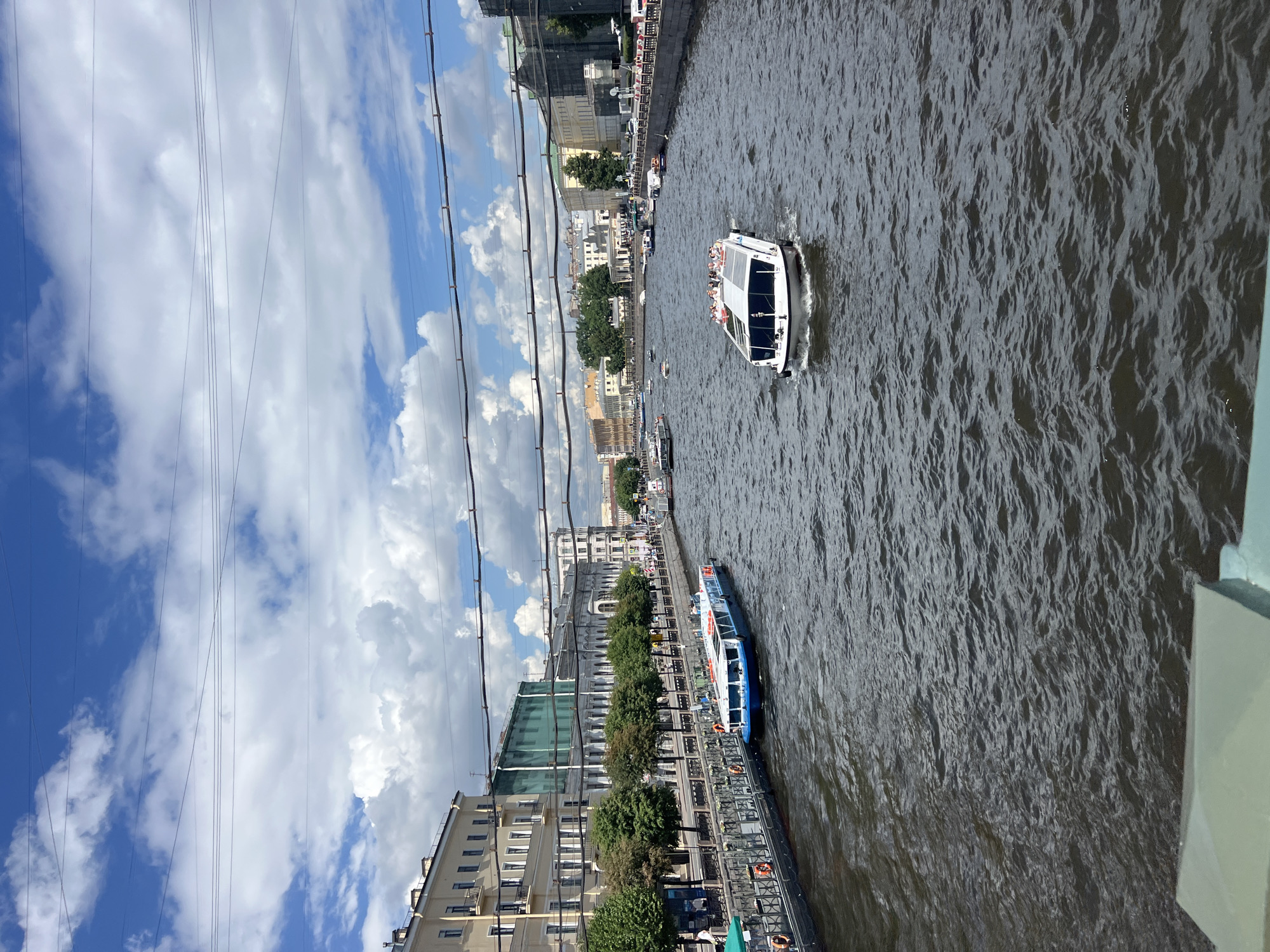 This image depicts a view of the Griboyedov Canal in Saint Petersburg, Russia, on a cloudy but bright day. The canal is filled with dark, rippling water, reflecting the sky and the surrounding architecture.
In the foreground and middle ground, several boats navigate the canal. A large, white passenger boat with a closed cabin and open-air seating at the top is moving away from the viewer, carrying a group of people. To the left, a blue and white passenger boat is docked along the embankment, with life preservers visible. Further down the canal, other smaller boats are visible.
The embankments on both sides of the canal are lined with buildings and trees. The left bank features a light-colored building with multiple floors and arched windows, followed by a row of green trees casting shadows on the canal. The right bank also shows a line of buildings and trees, with pedestrian activity on the walkway. Overhead, several thick wires crisscross the sky, likely for street lighting or power. The sky is a mix of blue patches and fluffy white clouds, indicating a dynamic weather pattern.