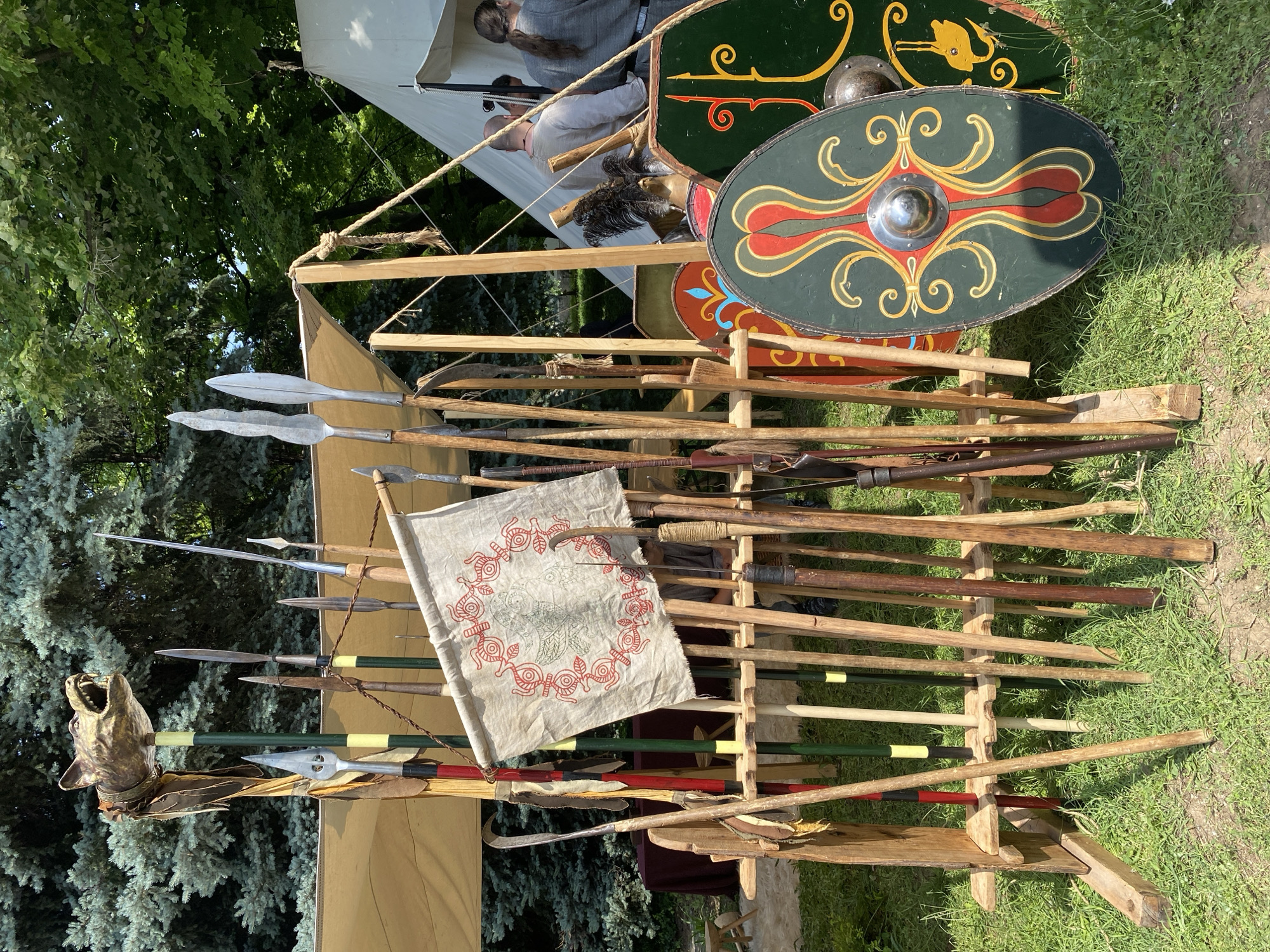 This image depicts an outdoor historical reenactment or exhibition display in the city of Pleven, Bulgaria. In the foreground, a wooden rack holds numerous spears, lances, and other polearms, varying in shaft color from natural wood to red and green, some with yellow bands. At the top center of the rack stands a prominent golden-colored effigy of a snarling wolf's head, mounted on a pole and adorned with leather strips. Below and to the left of the wolf's head, a light-colored fabric banner with a circular red patterned border and a central green abstract design hangs from a horizontal support.
To the right of the weapon display, several round and oval shields are visible, leaning on the grass. The most prominent is a dark green oval shield featuring a symmetrical central design of red and gold swirling patterns, with a metal boss in the middle. Behind it, a partially visible dark green shield shows a yellow stylized animal figure (possibly a rabbit or hare) and additional yellow scrollwork. Another red shield with blue and yellow motifs is partially visible further back. A feathered plume, possibly from a helmet, is also discernible among the shields.
In the background, under a large light-colored canvas canopy, a small group of people is visible. Two individuals, one with a ponytail, are seated or standing near a white canvas tent. Their attire appears casual. Beyond the canopy and tents, a dense natural backdrop of trees with both light green deciduous foliage and darker coniferous needles is present under clear daylight. The ground is covered with grass and some bare earth. A dark red tablecloth on a table is partially visible under the canopy on the left side of the frame.