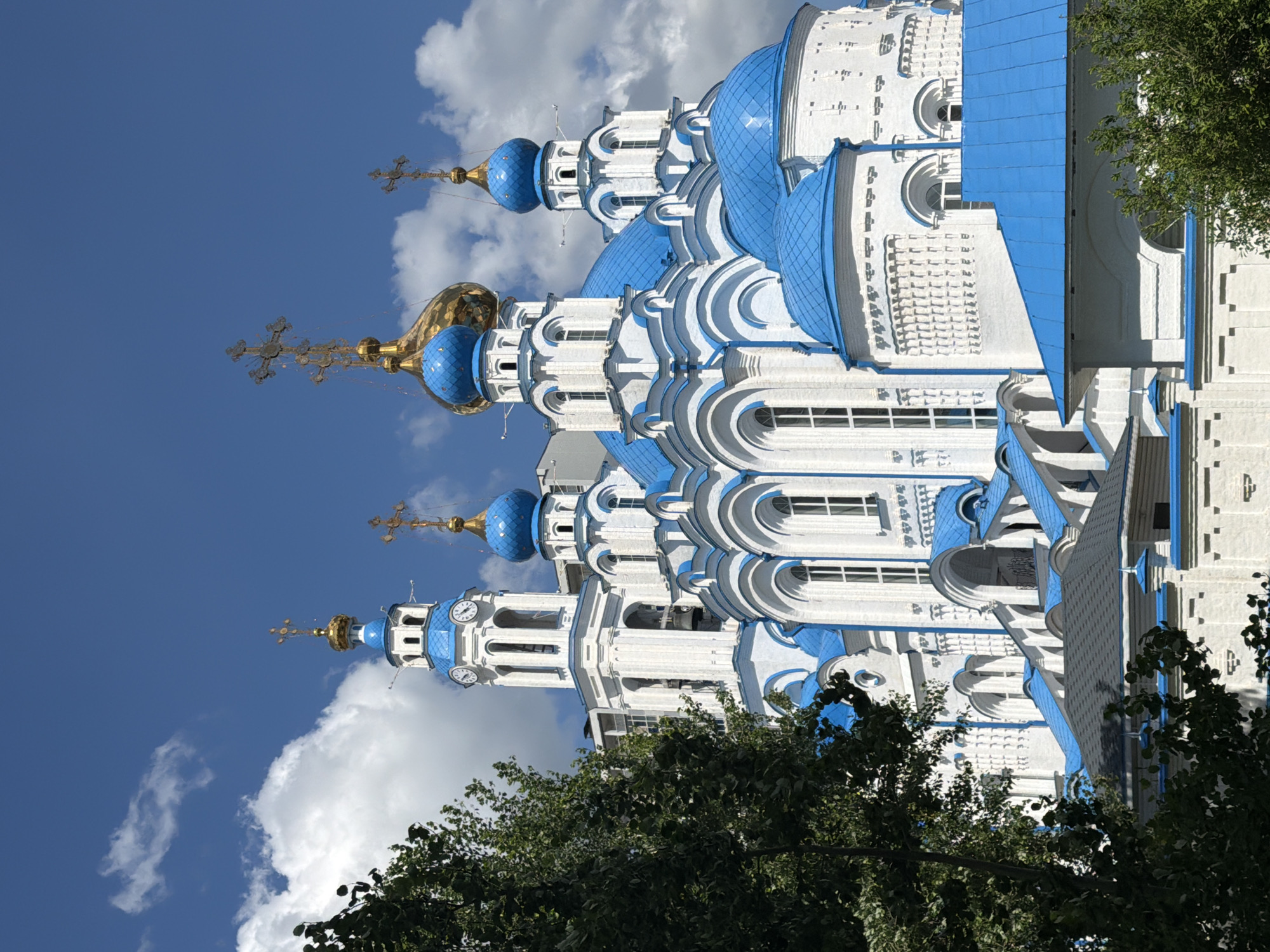 A white and blue orthodox church with multiple domes and a tall bell tower is visible against a bright blue sky with scattered white clouds. Green trees are in the foreground, partially obscuring the lower portion of the church. The church features intricate white architectural details, arched windows, and golden crosses atop its domes. The bell tower has clock faces. The scene appears to be a sunny day.