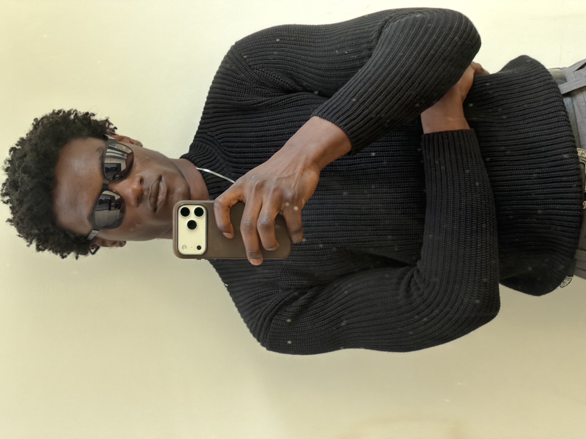 A dark-skinned man with curly hair is taking a selfie. He is wearing sunglasses and a black, ribbed sweater. He is holding a brown smartphone in his right hand, angled towards the viewer. A silver chain is visible around his neck. The background is a plain, light-colored wall.