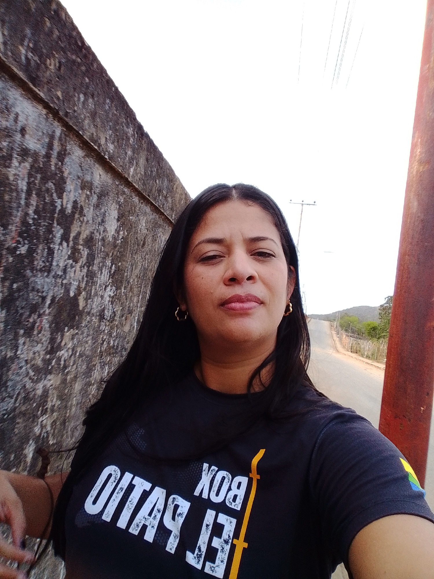 A woman is taking a selfie in Altagracia, Venezuela. She is standing next to a textured, rough concrete wall with a reddish-brown pole to her right. She has long, dark hair and is wearing a dark t-shirt with white and yellow lettering that reads "BOX" and "EL PATIO." She is also wearing gold hoop earrings. In the background, a paved road winds uphill with sparse vegetation and utility poles visible. The sky is bright white, indicating daytime.