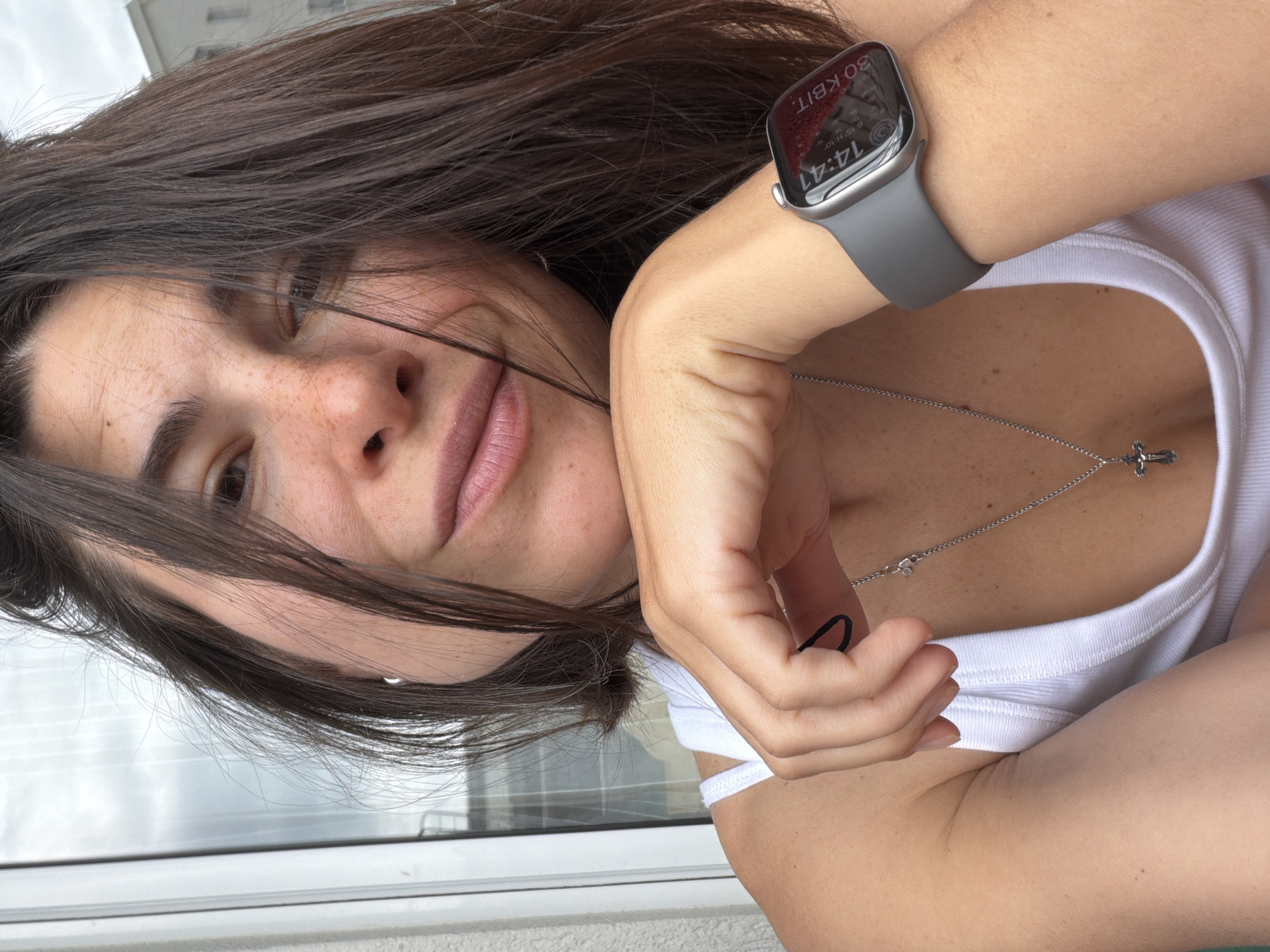The image is a close-up, tilted portrait of a woman, captured likely as a selfie, with a backdrop suggesting an outdoor setting in Budva, Montenegro. The woman has long, dark brown hair, part of which falls across her face. She has light skin, adorned with noticeable freckles on her nose and cheeks. Her eyes are partially visible, one looking towards the camera with a calm expression, while her lips are slightly parted, showing a natural or light pink color.
She is wearing a white ribbed tank top. On her left wrist, she sports a silver Apple Watch with a grey sport band. The watch display clearly shows the time as "14:41", along with Cyrillic text "30 KBіT." and smaller text "B 16 H:10". Around her neck, she wears a thin silver chain from which hangs a small, silver crucifix pendant. Her right hand is visible, holding a black elastic band, possibly a hair tie.
The background, though out of focus, reveals a cloudy, overcast sky and a hint of a building or balcony railing. The time displayed on the watch indicates it is early afternoon. The woman's expression is relaxed, suggesting a casual and comfortable moment.