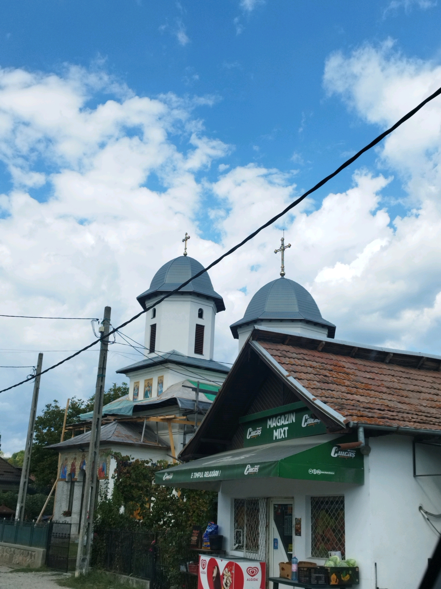 In Albeștii Ungureni, Romania, a white church with two grey-domed towers, each topped with a cross, stands against a bright blue sky with scattered white clouds. Power lines are strung across the foreground, intersecting with the view of the church. To the right of the church, a small white building with a brown tiled roof houses a "MAGAZIN MIXT" (Mixed Store). The store has a green awning with logos, and its windows display merchandise, including vegetables and what appears to be frozen treats. A black metal fence lines the front of the property. The scene is set during the daytime, under clear weather conditions.