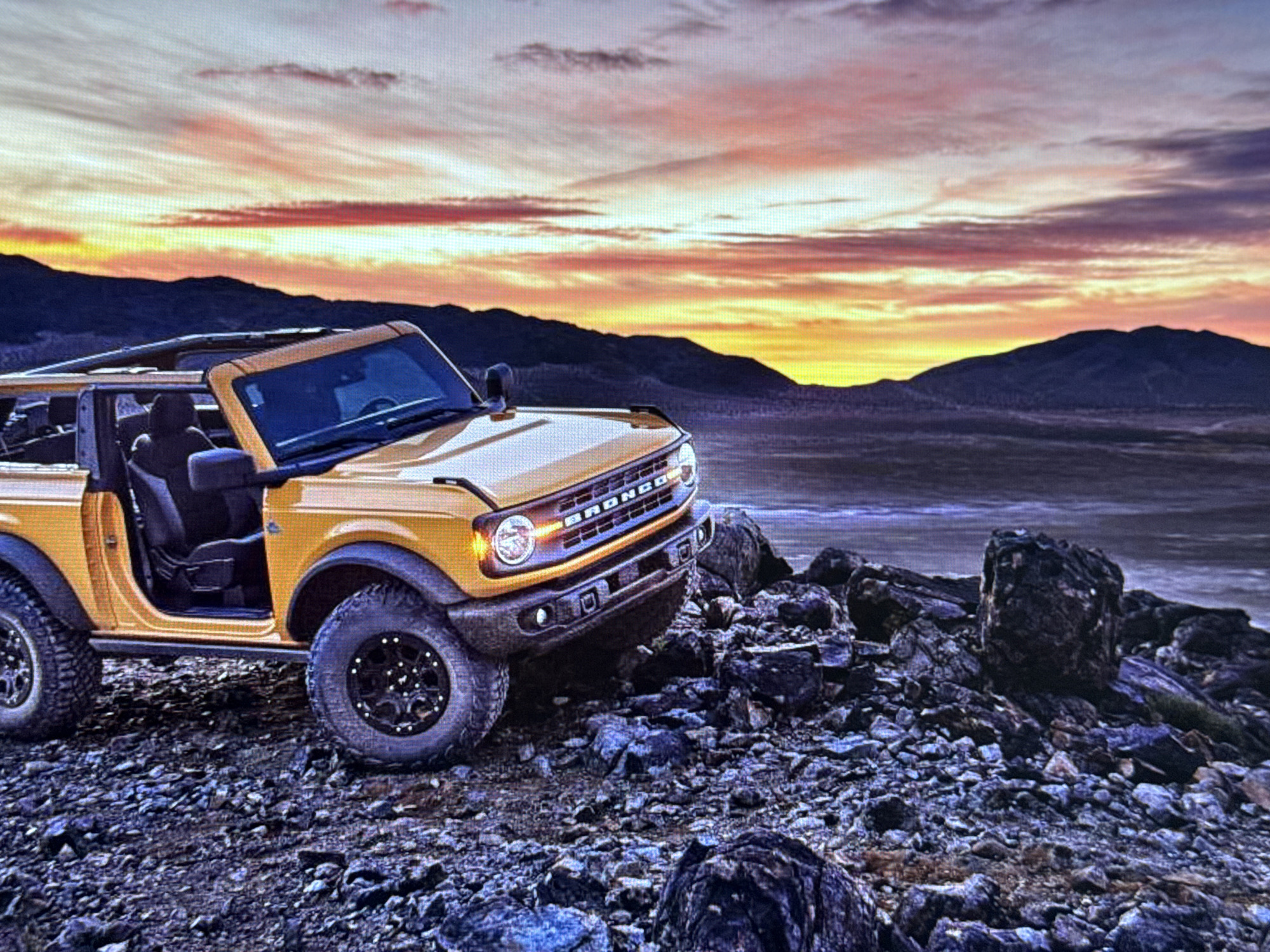 A yellow Ford Bronco is parked on a rocky, uneven terrain, angled towards the left of the frame. The vehicle has its driver's side door open and the convertible top removed. The sky above is filled with a dramatic sunset, featuring vibrant hues of orange, pink, and yellow. In the background, dark, silhouetted mountains rise against the colorful sky, and a vast, barren landscape stretches out before them. The foreground is dominated by large and small rocks of varying shades of gray and brown. There are no people visible in the image. The scene does not depict any recognizable landmarks or features of Baku, Azerbaijan.