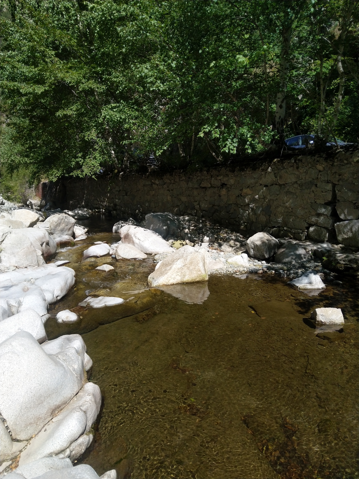 The image depicts a shallow, clear stream flowing over rocks and pebbles. Large, smooth, white boulders line the left bank of the stream, with smaller rocks and gravel visible on the right bank. A stone wall runs along the upper bank, overgrown with vegetation, including trees with dense green foliage that casts shadows on the wall and the stream. A dark-colored car is partially visible among the trees on the far bank. The water is shallow, allowing the rocky bottom to be seen, and exhibits ripples and reflections of the surrounding foliage and sunlight. The scene appears to be a natural setting, possibly a park or a natural reserve, and is located in Garliano, Bulgaria.
