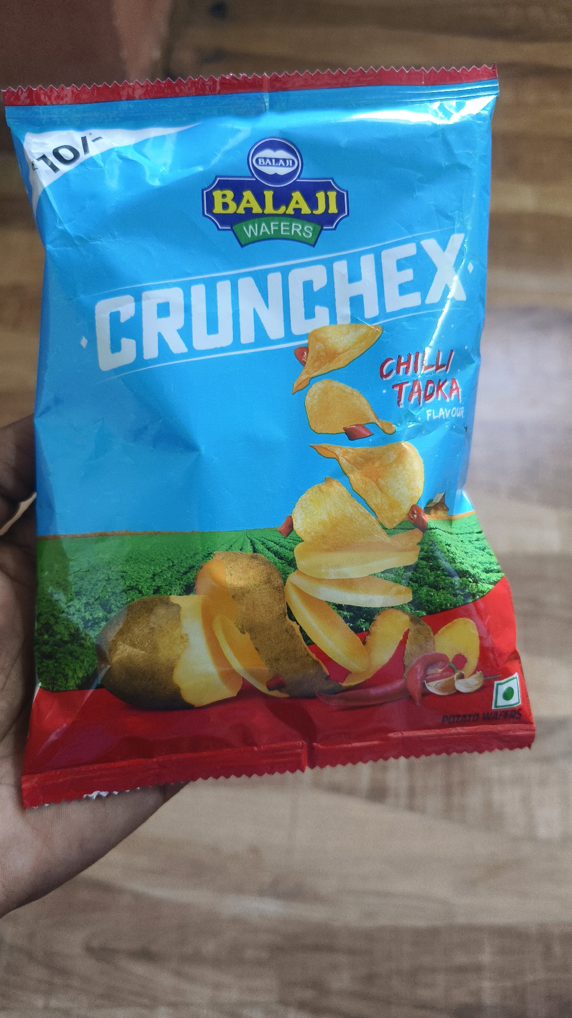 The image displays a bag of "Balaji Wafers" brand potato chips, specifically the "Chilli Tadka" flavor. The bag is primarily bright blue with red accents. The brand logo, featuring the name "Balaji" in yellow lettering within a blue crest, is prominently displayed at the top. Below the logo, the product name "CRUNCHEX" is written in large, white, sans-serif capital letters. Scattered around the product name are images of potato chips, whole potatoes, and chili peppers. The background depicts a green field, suggesting the origin of the potatoes. The bottom of the bag is red and has a zigzag edge, typical of chip packaging. A small green square with a white circle inside, indicating a vegetarian product, is visible on the lower right. A hand is holding the bag, obscuring the lower left portion. The price, "10/-", is printed on the upper left of the bag. The scene is likely set indoors, with a wooden surface visible in the background, suggesting a casual setting. There is no discernible information to suggest the location is specifically in Belagavi, India; it is a generic product image.