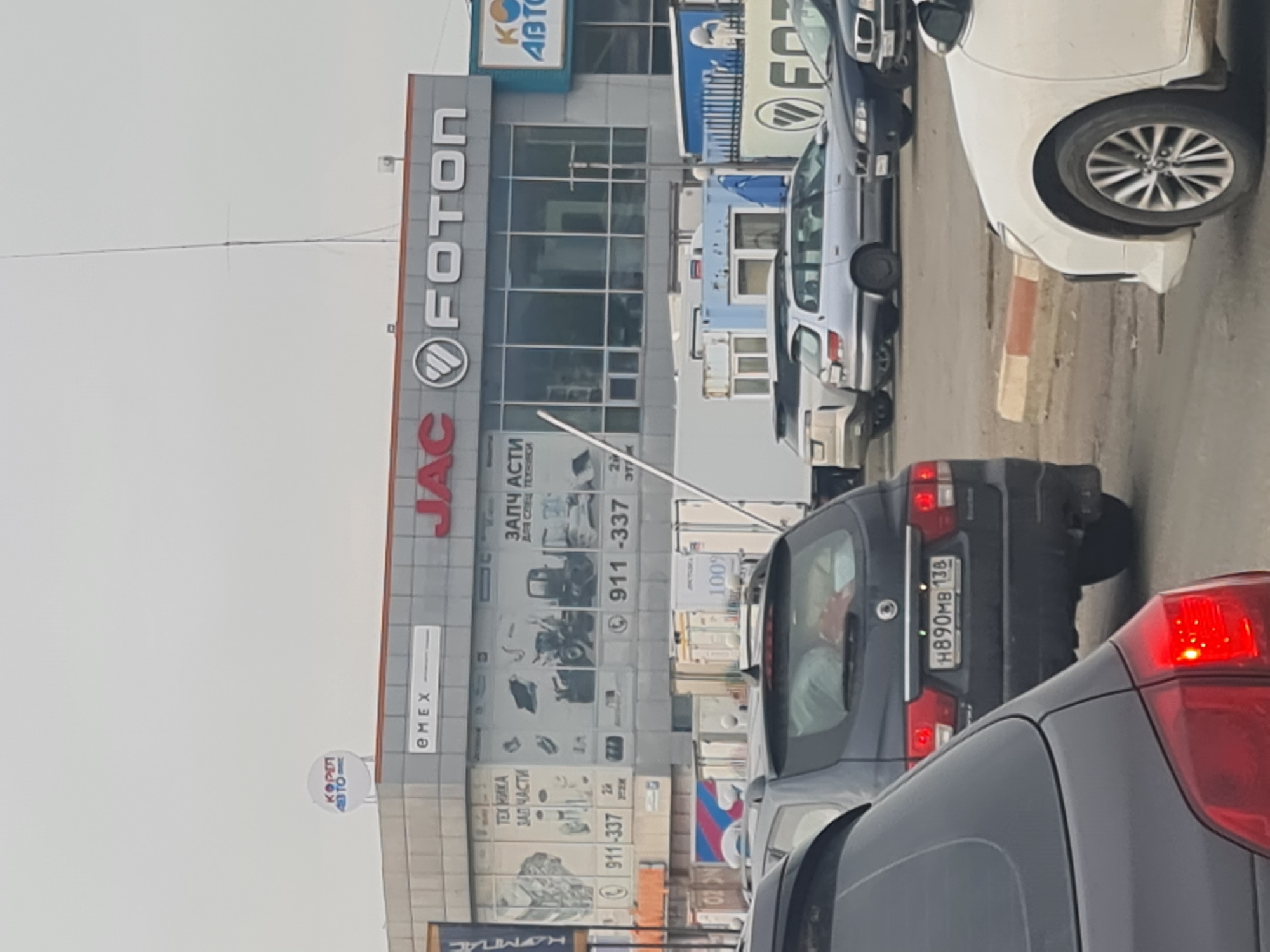 A street view in Irkutsk, Russia, shows a building with signs for "EMEX", "JAC", and "FOTON". The building appears to be a commercial establishment selling vehicle parts and possibly vehicles. Several cars are visible in the foreground, suggesting traffic or a parking lot. The overall scene depicts an urban commercial area with typical infrastructure and vehicles.