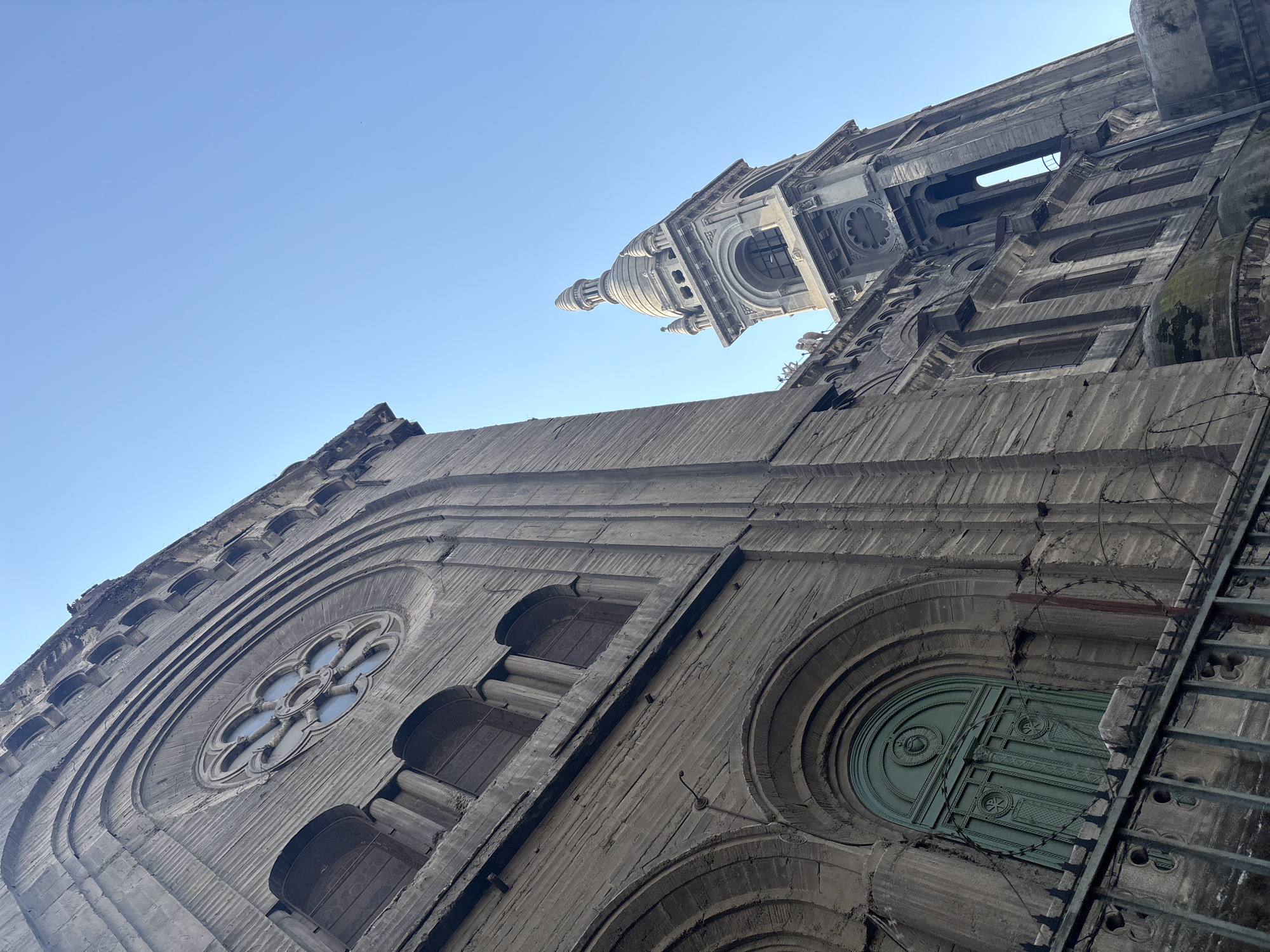 This is a low-angle outdoor photograph of the Church of San Vicente Ferrer in Santiago, Chile. The church is made of a light-colored stone or concrete material and features Gothic architectural elements. In the foreground, a portion of the church's facade is visible, including arched windows and a large, ornate green double door. A metal fence with barbed wire is positioned in front of the door. To the right of the foreground section, a tall bell tower with a conical roof rises against a clear blue sky. The bell tower has multiple levels with arched openings and decorative stonework. The overall scene suggests an aged building with a sense of historical significance. The sky is clear and bright, indicating daytime. There are no people visible in the image.