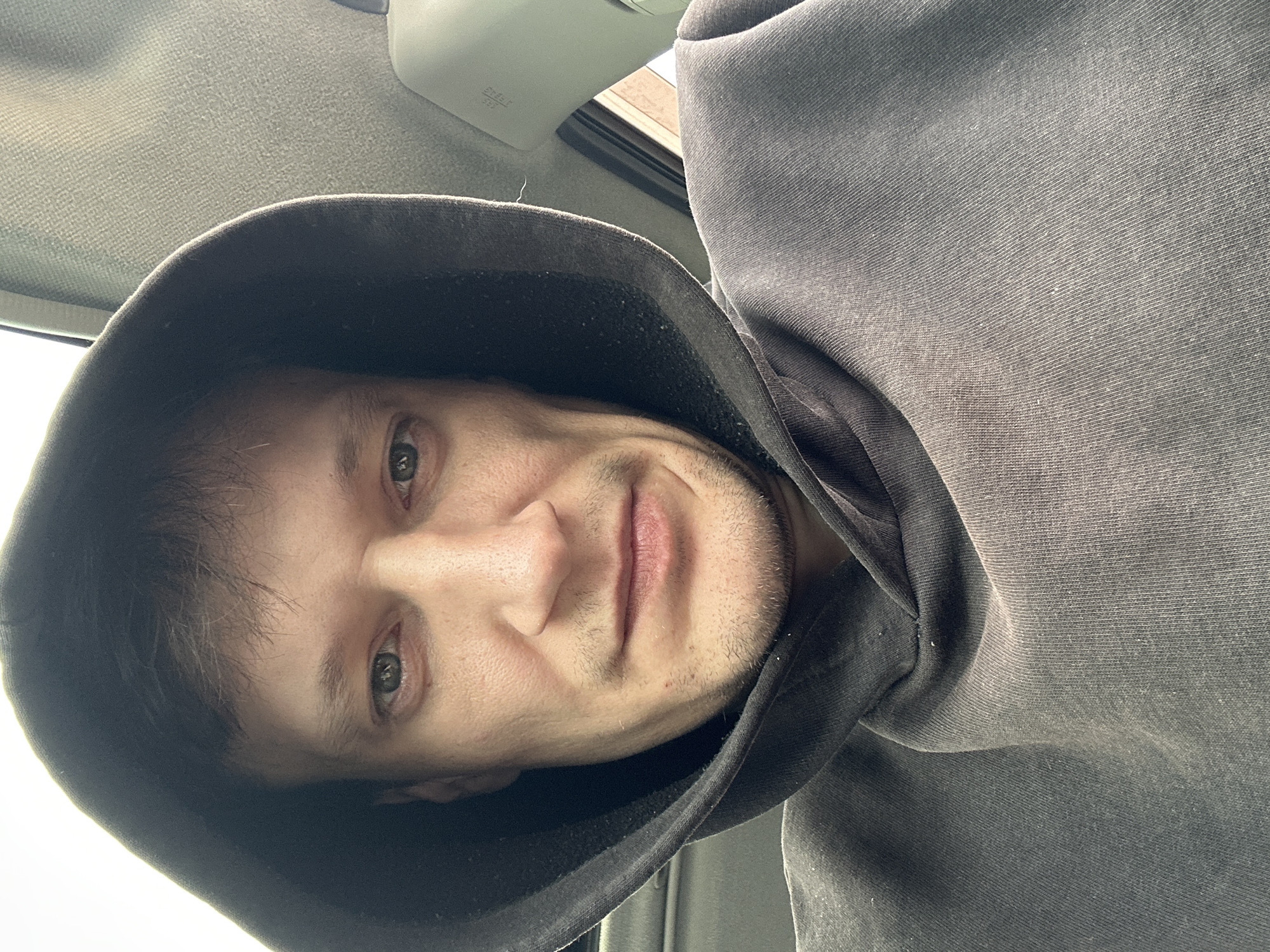 This is a close-up, selfie-style, indoor photograph of a person wearing a dark gray hooded sweatshirt. The individual has light skin, short dark hair, and light green eyes. Their face is looking directly at the camera, with a neutral expression. The background appears to be the interior of a vehicle, with a textured light gray material on the right side, possibly a car headliner or door panel. There is also a faint hint of a window with light filtering through. The image quality is slightly grainy, suggesting it was taken with a mobile phone camera. The location is not specified in the image data or its context, and therefore cannot be determined to be in Makiv, Ukraine.