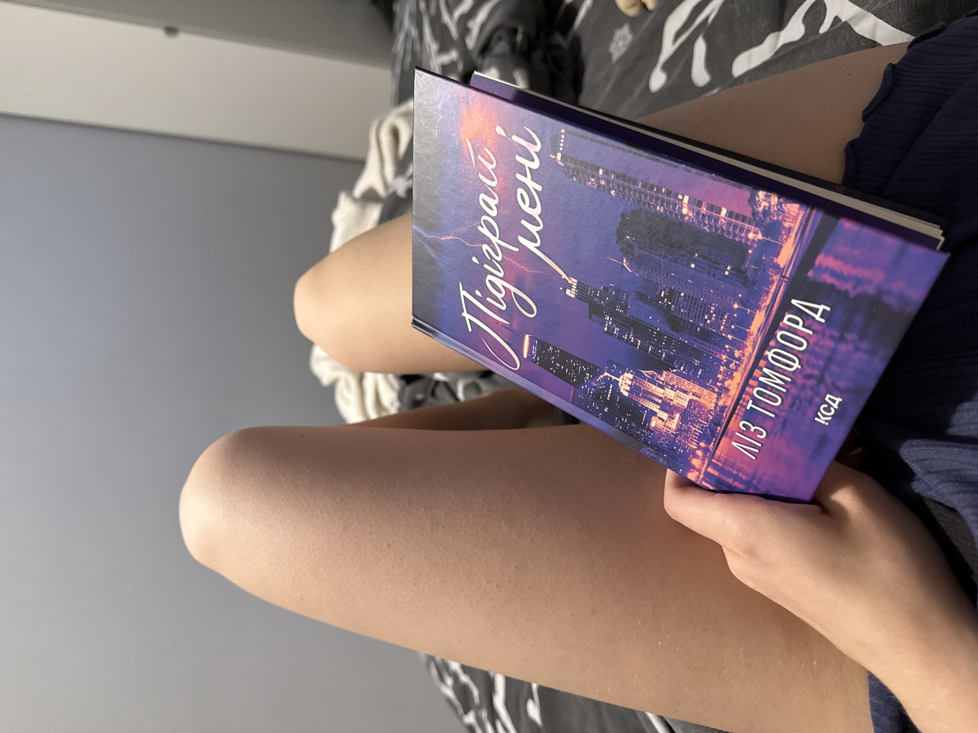 A book lies open on a person's lap. The book's cover features a vibrant depiction of a city skyline at night, bathed in purple and orange hues, with streaks of lightning illuminating the sky. The title, in elegant cursive, is visible on the cover, as is "ЛІЗ ТОМФОРД" and "КСД" which are likely author and publisher information. The person's legs are crossed, and they are wearing dark blue clothing. The background consists of a bed with patterned bedding and a plain gray wall. The scene suggests a moment of quiet relaxation, perhaps indoors.