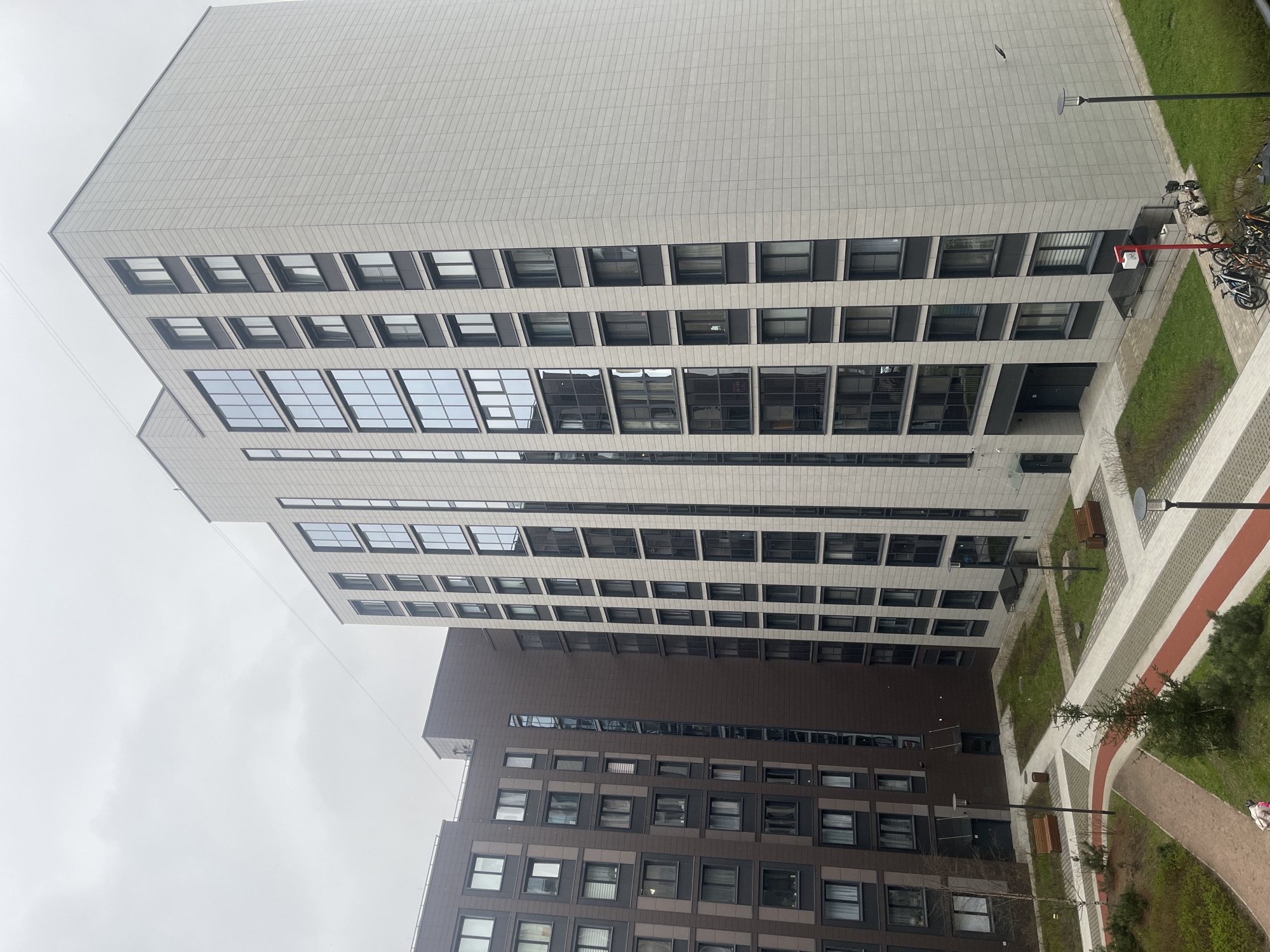 An elevated view captures a modern urban scene featuring two multi-story buildings and an intervening courtyard under an overcast sky.
The prominent building on the left is tall, with a light gray or beige facade composed of a grid of rectangular panels. It features numerous windows, some arranged in large, multi-pane sections, and others as individual rectangular openings, all framed in dark material.
To the right, a second, slightly lower building is visible, constructed with a dark brown facade, also displaying a grid pattern and dark-framed windows.
The courtyard, situated between and in front of the buildings, is a mix of paved areas and greenery. Light gray tiled pathways are interspersed with darker gray sections and a striking red stripe, creating a geometric design. Patches of green grass and a few small trees are planted within these paved areas.
Several bicycles are parked near an entrance of the light-colored building, accompanied by a red bollard or stand. Multiple lampposts illuminate the courtyard. The sky is entirely overcast and grey, indicating a cloudy daytime setting. A thin cable or wire spans the upper portion of the frame against the sky. A faint, light-colored object, possibly a person or bag, is discernible on a path in the lower right of the courtyard.
No direct human activity is clearly visible, and no text is discernible within the image.