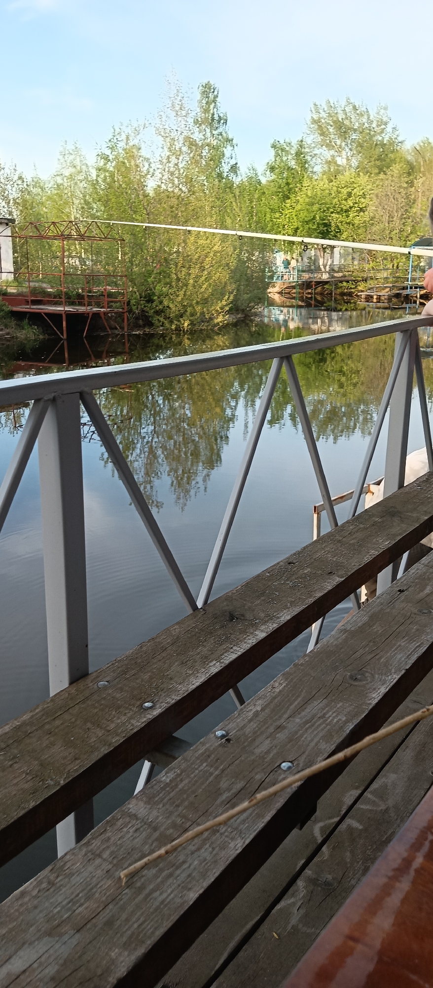 The image depicts a view from a wooden bench with a metal railing overlooking a body of water, possibly a lake or river. Across the water are platforms or small structures nestled among trees and foliage. The reflections of the trees are visible on the water's surface. The scene appears to be in a natural or park-like setting, with a clear sky visible in the upper portion of the image. The benches and railing are in the foreground, providing a sense of perspective and depth to the landscape.