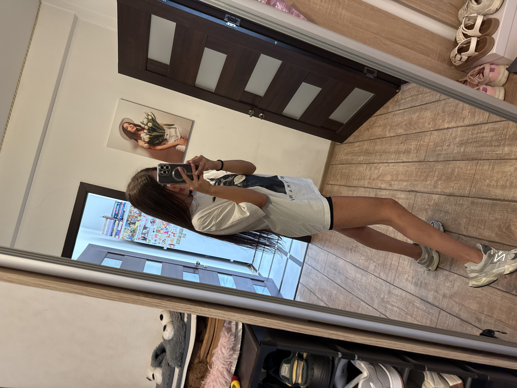 A young woman with long, dark hair is taking a mirror selfie in a room. She is wearing an oversized white t-shirt and black shorts, with light gray sneakers. In the reflection of the mirror, a painting of a woman holding tulips hangs on the wall. To the right, there is a dark wooden door with glass panes. To the left, a shoe rack is visible, holding several pairs of shoes and slippers. The room has wooden flooring and the lighting suggests it is daytime. The setting appears to be a residential interior, likely a bedroom or a hallway, in Chișinău, Moldova.
