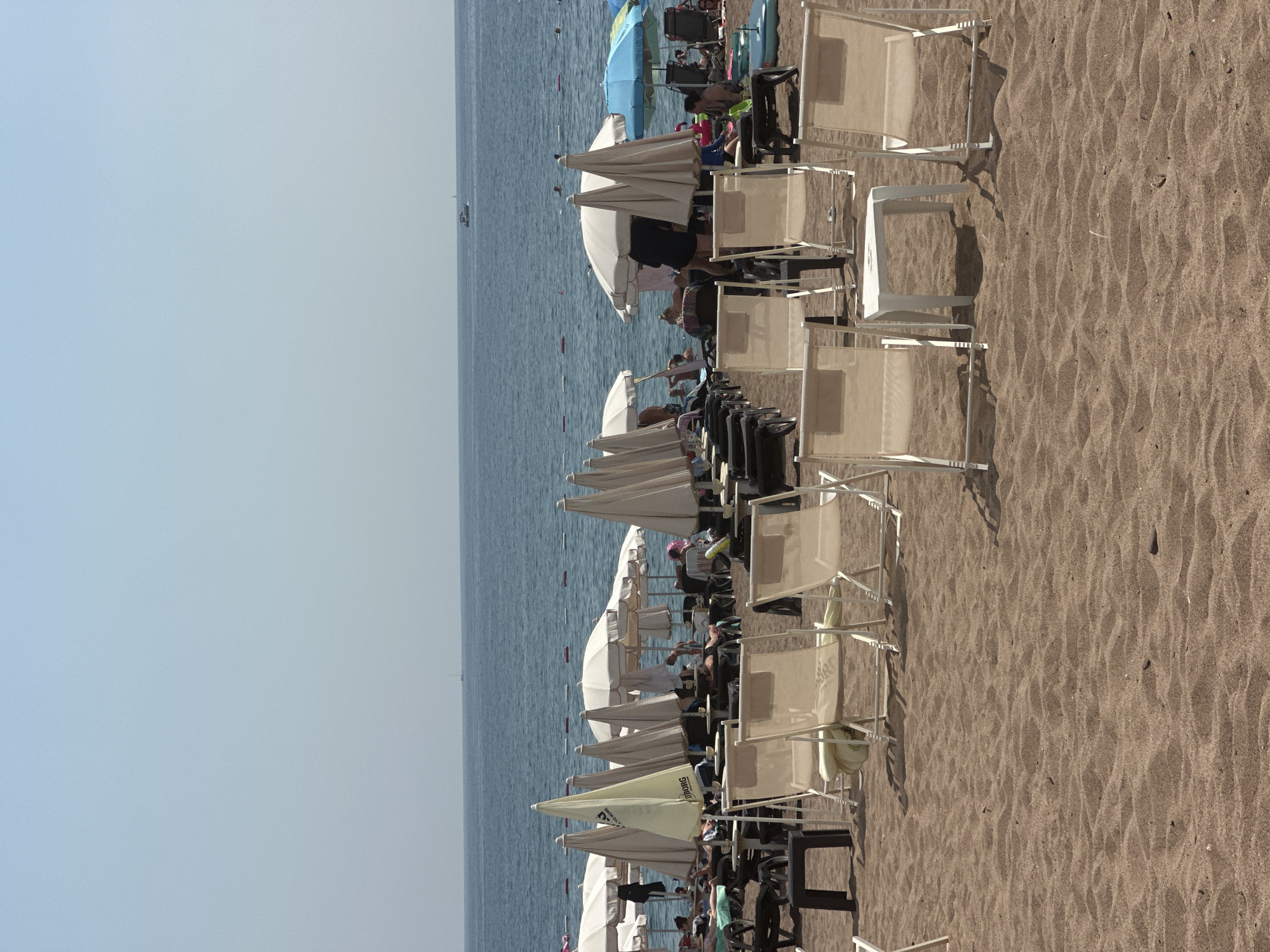 The image depicts a crowded beach in Boreti, Montenegro. In the foreground, several empty, beige deck chairs with white frames are arranged on the sandy beach. A small, white, cube-shaped table is positioned between two of these chairs. The middle ground of the image is filled with more deck chairs and a variety of closed and open umbrellas, mostly in white and beige tones. Some people are visible relaxing on lounge chairs or walking among the umbrellas. Further back, in the background, the blue sea stretches out, with a few buoys marking a swimming area. A small white boat is visible on the horizon, and a distant, possibly larger, sailboat can be seen. The sky is a clear, pale blue. The overall impression is a sunny, active beach scene.