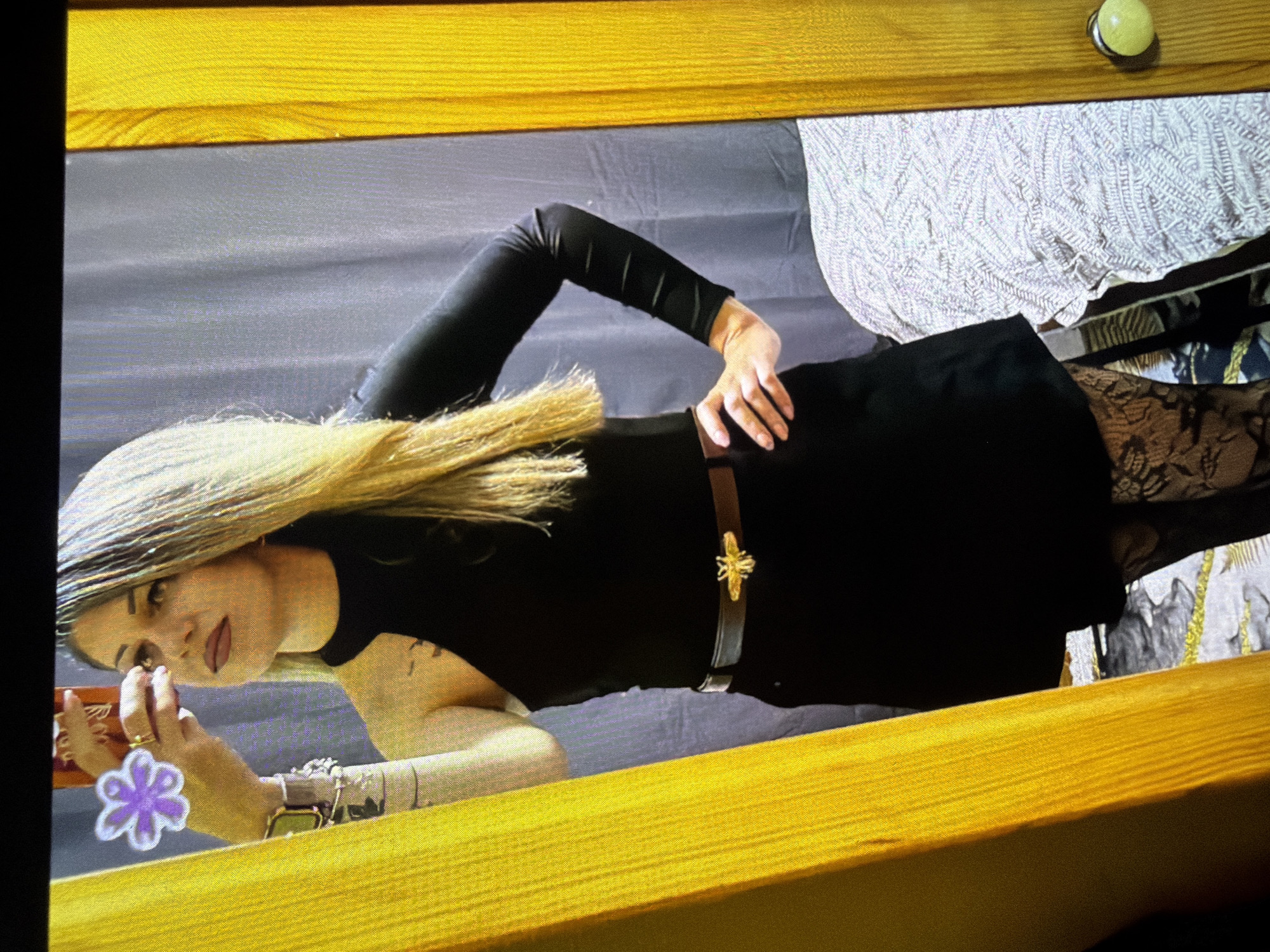 A woman with blonde hair and fair skin is lying on her side on a surface covered with a gray cloth. She is wearing a black dress cinched at the waist with a brown belt that has a gold bee buckle. She has on long black sleeves, patterned stockings, and a variety of jewelry on her left wrist. Her left hand holds a rectangular object, and a purple flower adorns her finger. The surrounding environment features yellow wooden structures. The location in Dryanovo, Bulgaria, is not discernible from this image alone.