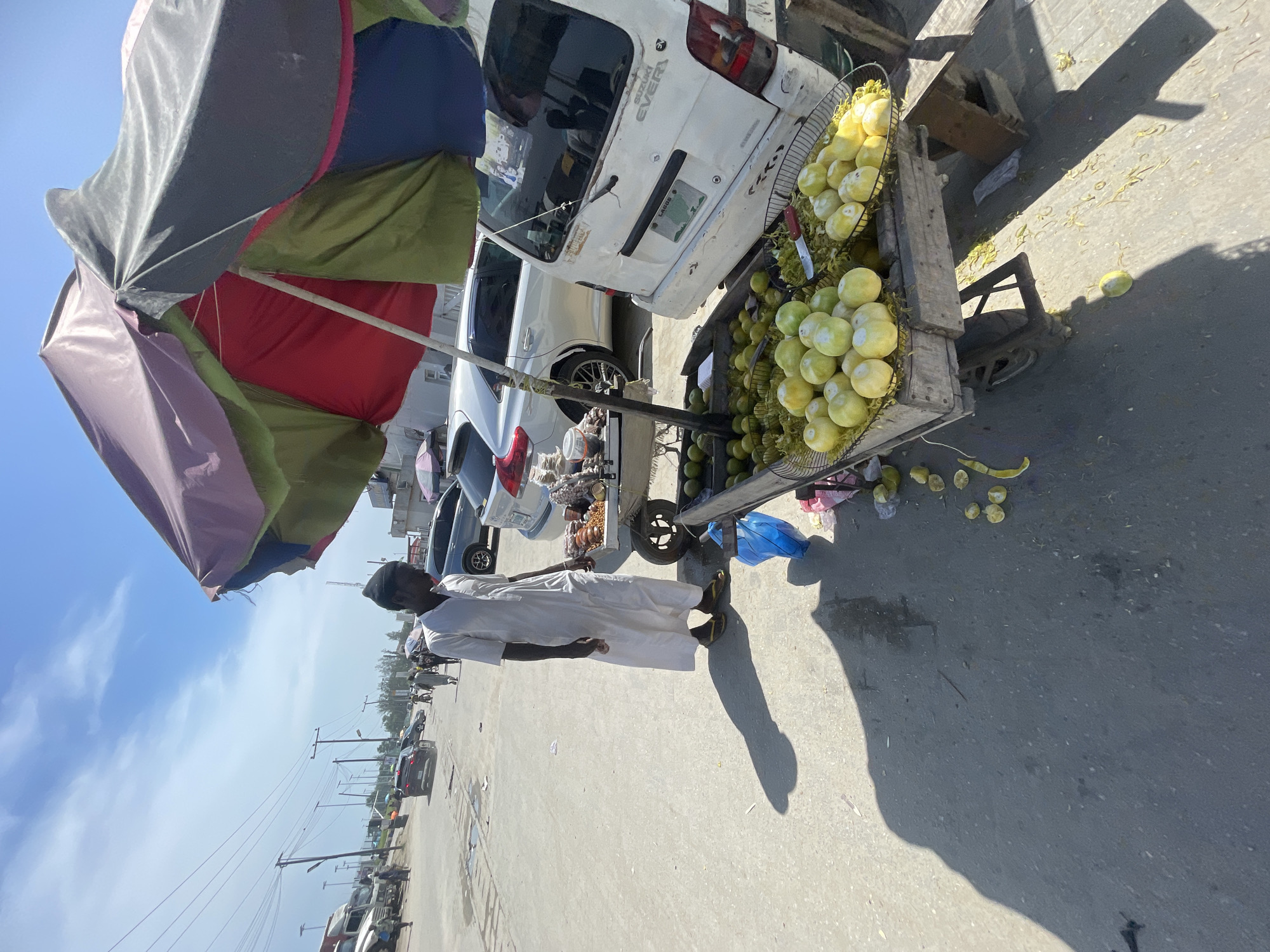 This image captures a bright, sunny street scene in Lagos, Nigeria, focusing on a street vendor and his fruit stall. A man, dressed in a white tunic and a dark cap, stands alongside a wooden cart. He appears to be the vendor, attending to his merchandise.
The cart is shaded by a large, multicolored umbrella featuring panels of gray, red, green, and light purple. The cart itself is laden with light green and yellow fruits, likely guavas, displayed in metal baskets. A knife with a red handle is visible among the fruits. Scattered fruit peels and small pieces of fruit are on the asphalt ground near the cart, along with a blue plastic bag.
Behind the cart, several white cars are parked. One prominent vehicle is a white Suzuki Every van, with "SUZUKI EVERY" visible on its rear and a license plate clearly displaying "LAGOS," confirming the geographical context. Other vehicles and buildings line the street in the background.
The time of day is indicated by the strong, direct sunlight and the long, distinct shadows cast by the man and the cart on the ground, suggesting midday or early afternoon. The sky is a clear blue with a few wispy clouds. Power lines and utility poles are visible in the distance, characteristic of an urban or semi-urban environment. The scene depicts a common roadside vending activity in Lagos.