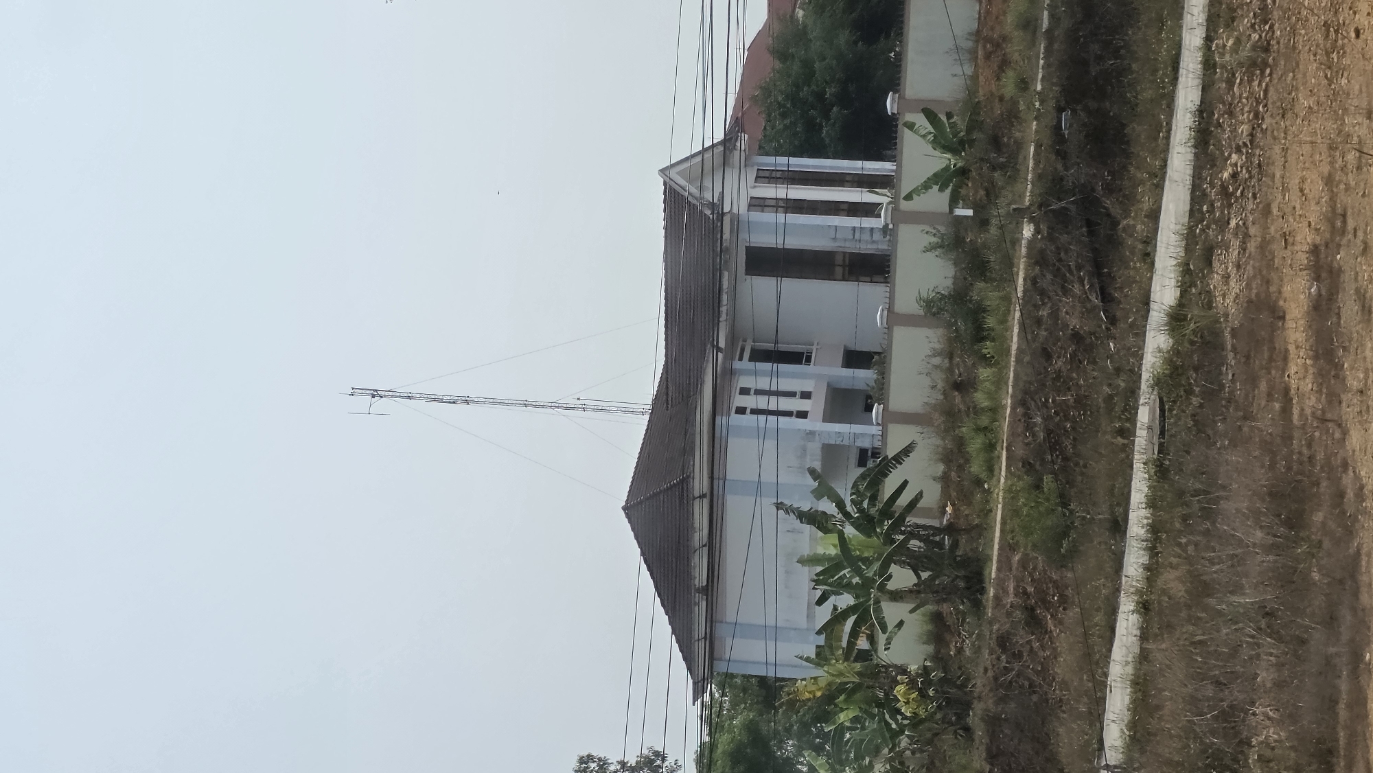 The image shows a two-story building with a brown roof in Tân Cảnh, Vietnam. A tall antenna rises from the roof. Power lines crisscross the sky in front of the building. Banana trees grow in the yard. A concrete wall runs along the edge of the property, separating it from a dirt embankment. The sky is overcast.
