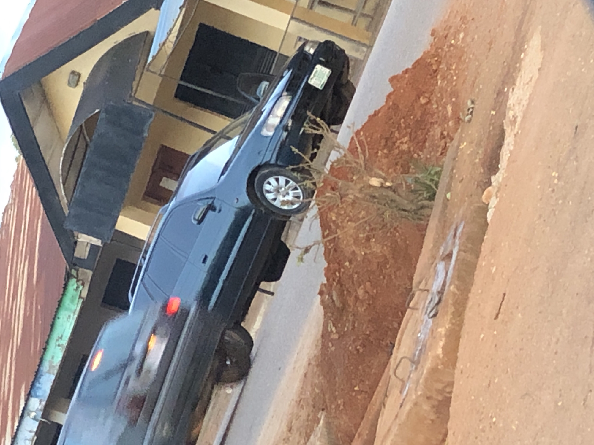 The image presents a tilted view of a street scene in Kaduna, Kaduna State, Nigeria, featuring two dark-colored vehicles and several buildings under a light blue sky.
In the immediate foreground, a significant pile of reddish-brown dirt occupies the lower left, extending partially across the bottom. A concrete slab, possibly a manhole cover or drainage component, is visible at the bottom center with two embedded metal handles and white residue. Scattered small, greyish objects, possibly stones or debris, are on the dirt and ground to the right of the pile. Sparse, dry vegetation protrudes from the dirt embankment.
The midground features a paved road, likely asphalt, running diagonally from left to right. A dark-colored (appears dark green or black) station wagon or large sedan is prominently parked or driving on the right side of the road, facing towards the right. Its front passenger-side wheel displays a silver multi-spoke alloy rim. The front bumper holds a Nigerian license plate that reads "KADUNA KADUNA 006G05". The vehicle has a roof rack. Behind this vehicle, to its left, another dark-colored vehicle is partially visible, exhibiting motion blur. Its red taillights are illuminated, along with an orange indicator or side marker light.
The background consists of several buildings. The most prominent structure on the right is a two-story building with a light yellow/cream facade and a reddish-brown corrugated metal roof. A distinctive dark-colored, angular, cantilevered structure extends from its upper facade. An arched opening is visible above this structure, and a dark window or entrance is below it. To the left, another building is partially visible, also with a reddish-brown corrugated roof and a weathered light blue-green fascia board. The upper portion of the frame shows a clear, bright sky. No individuals are visible in the scene.